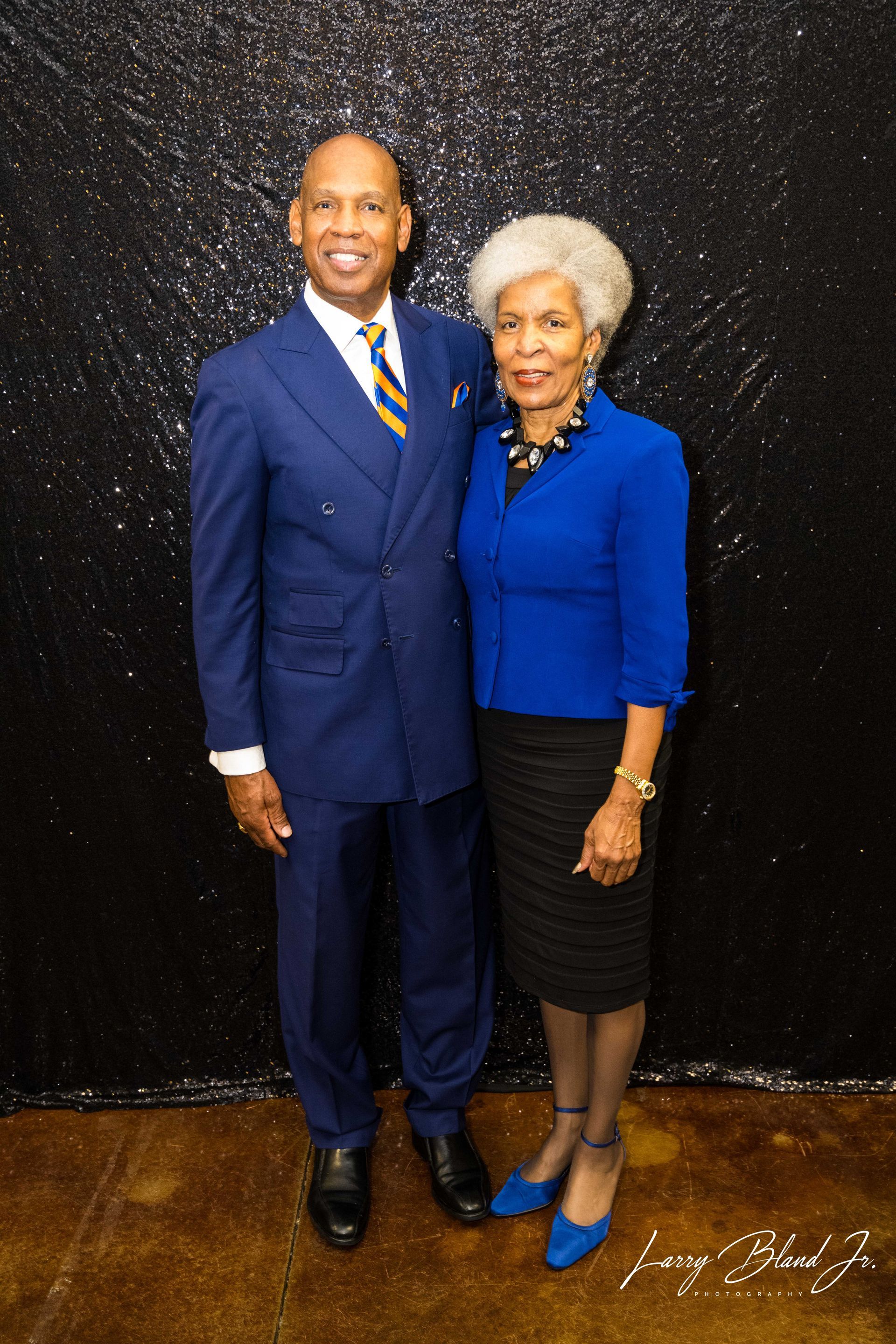 Man in blue suit and woman in blue jacket stand next to each other in front of a black backdrop.