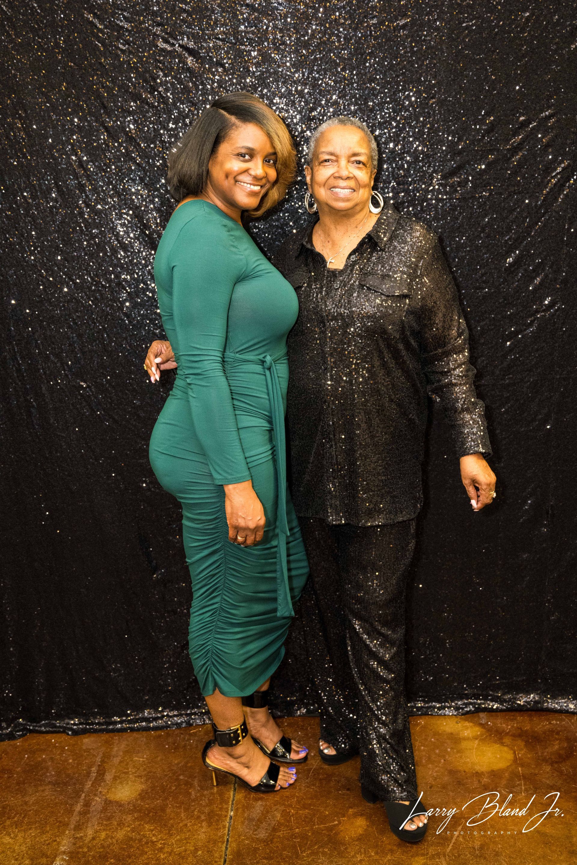 Woman in green dress and woman in black sequin outfit posing in front of a glittery black backdrop.