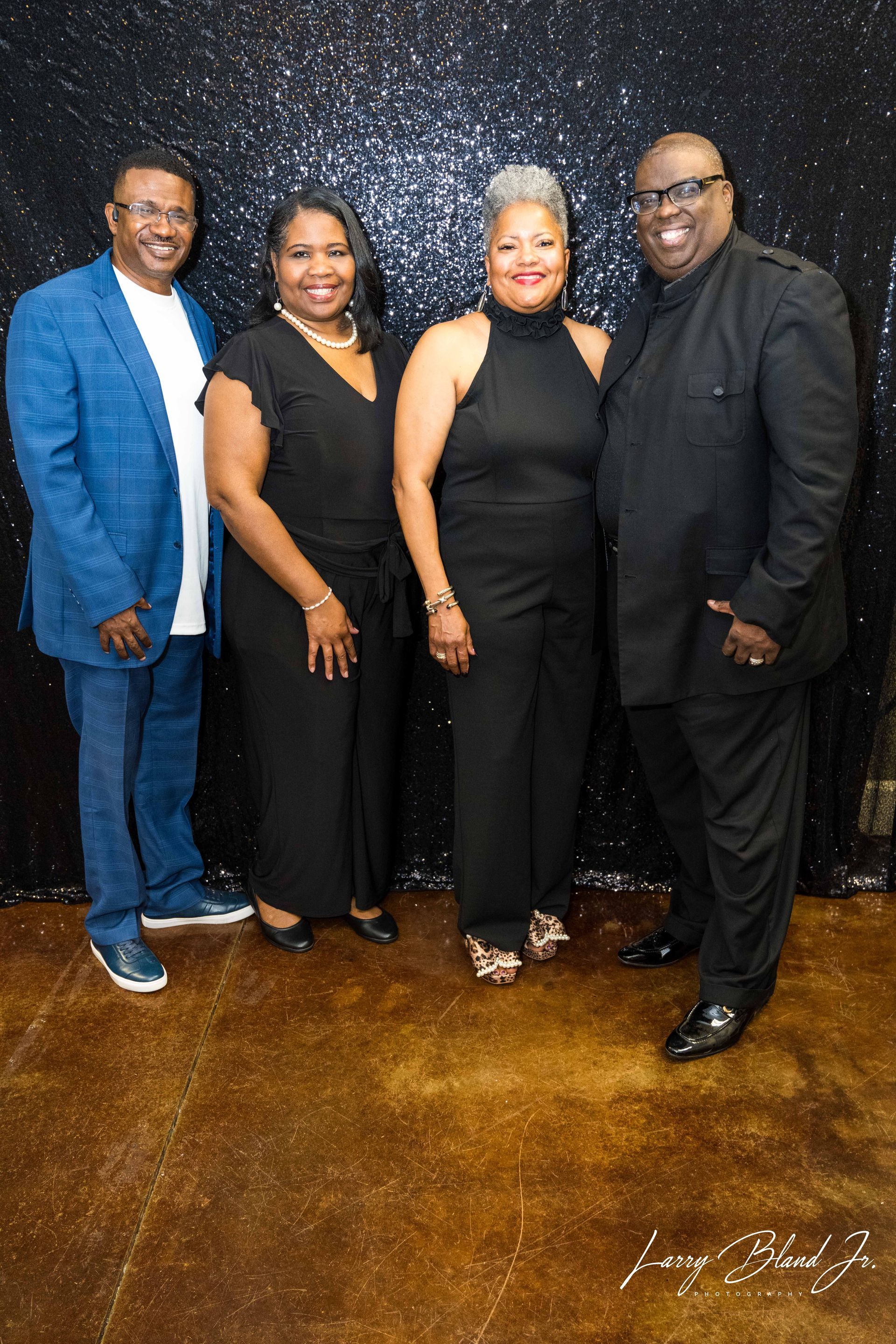 Four people posing in front of a sparkly black backdrop. Two women and two men wearing formal attire.