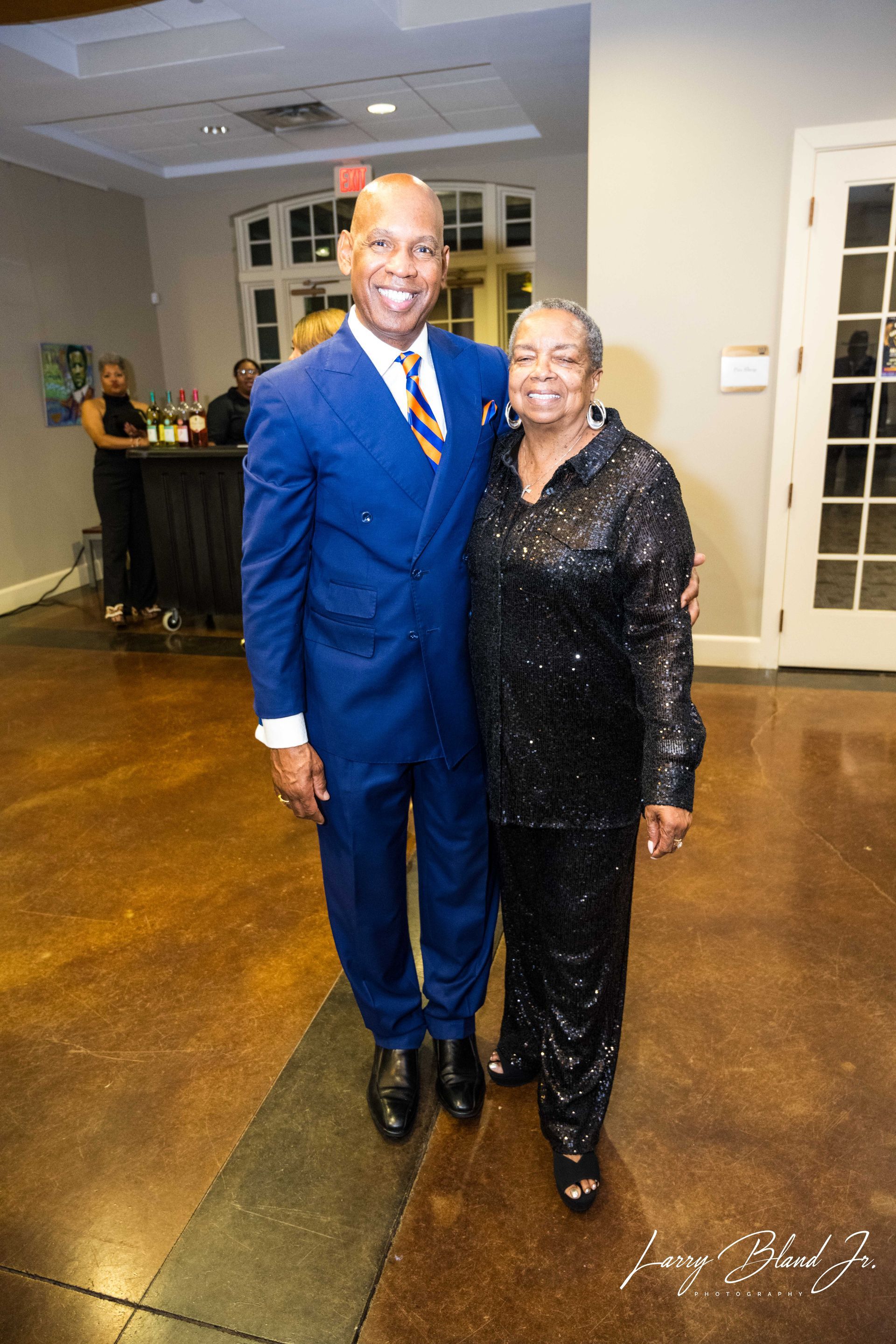 Man in blue suit and woman in black sequined outfit pose together indoors, smiling.
