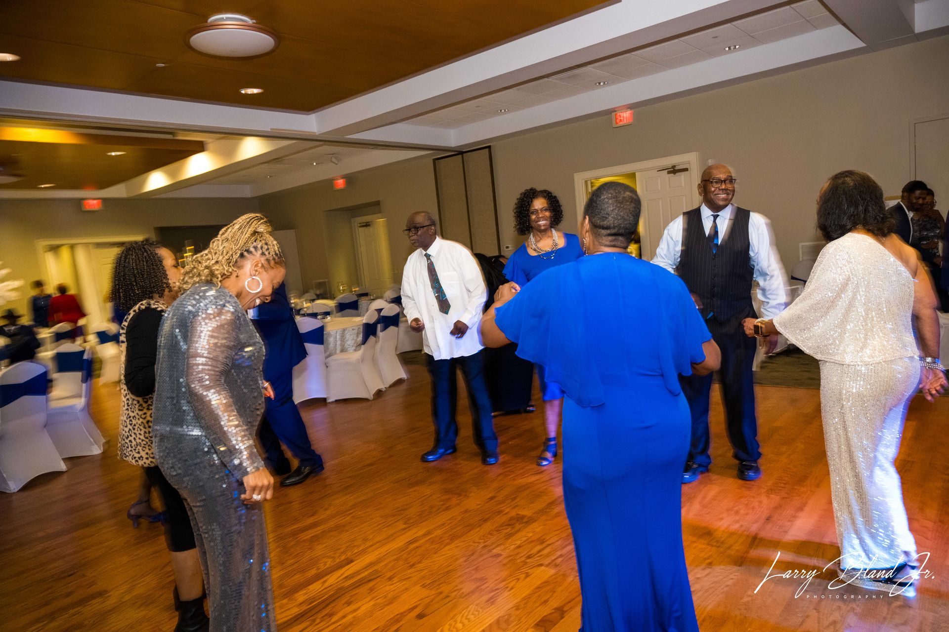 People dancing on a wooden floor in a banquet hall. Some wear blue and silver outfits.