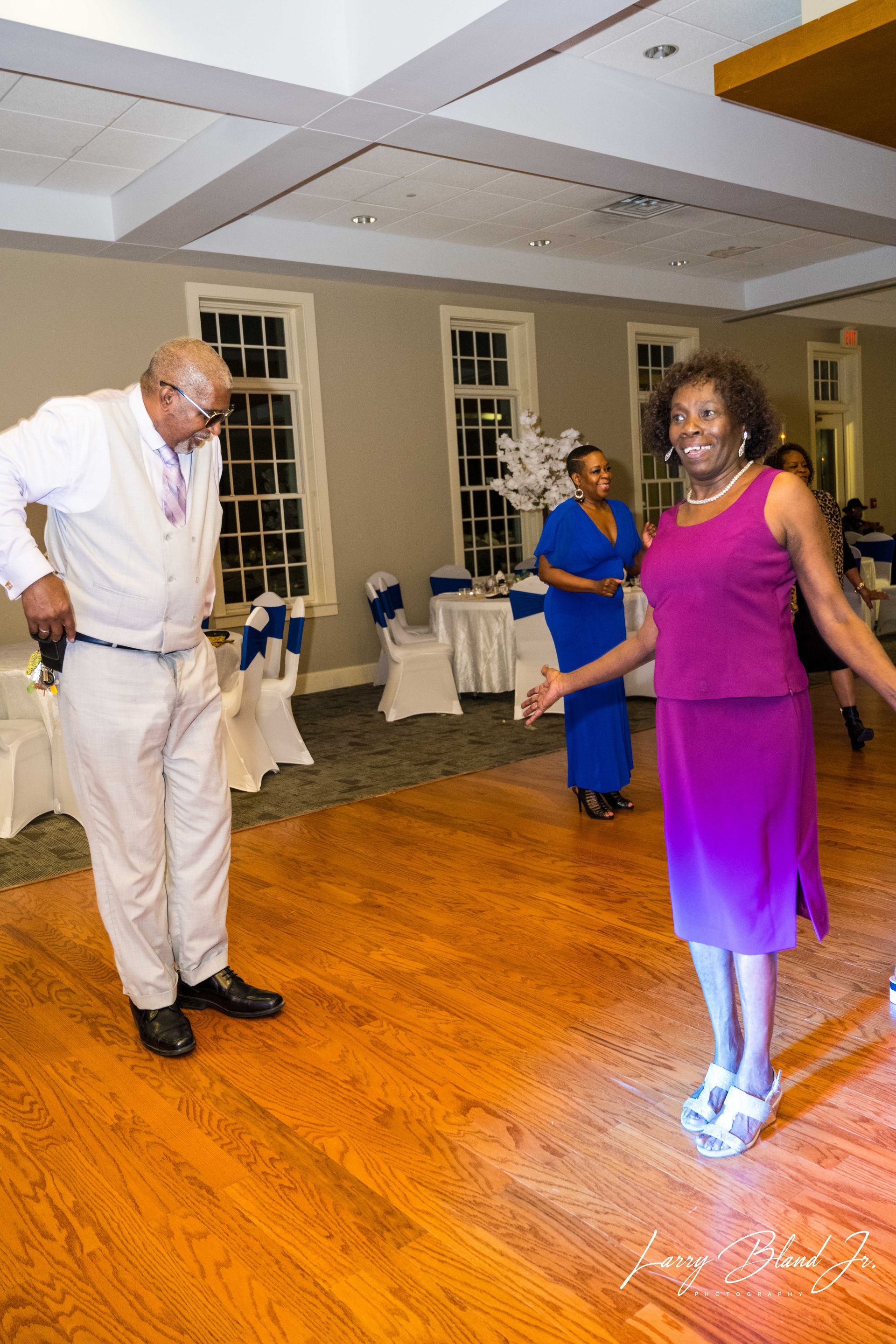 Man and woman dancing on a wooden floor in a banquet hall; other people and tables are in the background.