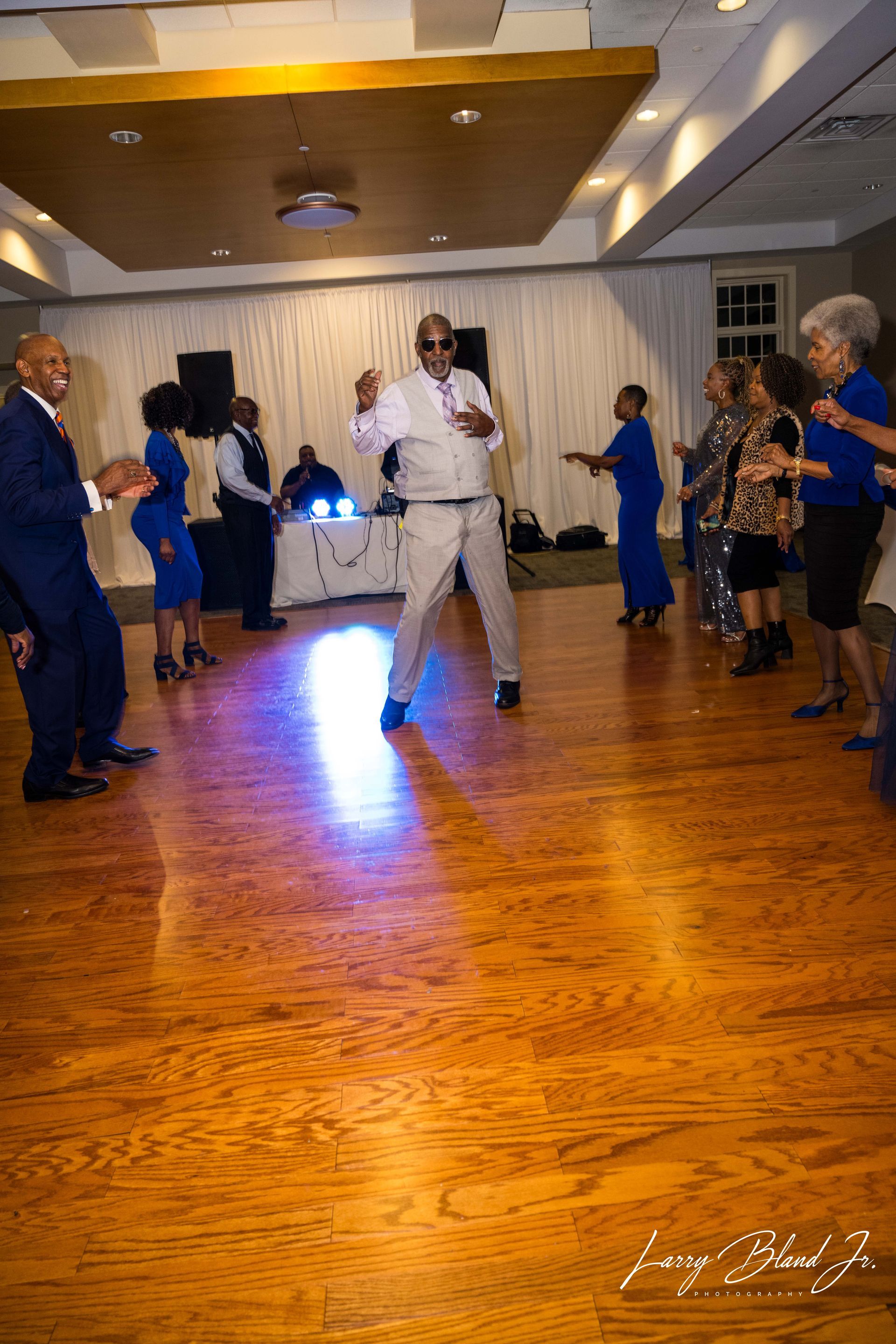 People dancing on a wooden floor in a room. A man in sunglasses dances in the center, others look on.