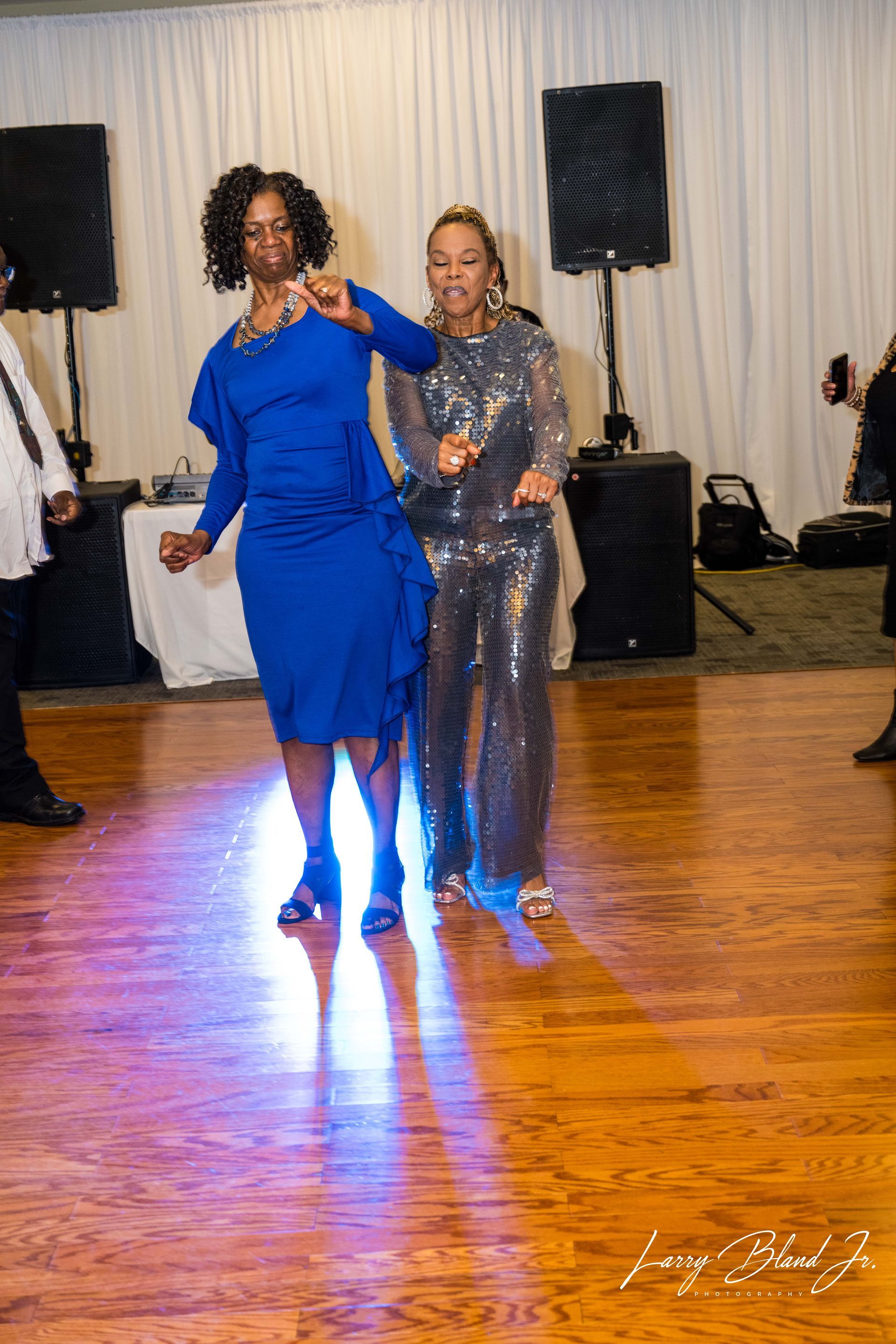 Two women dancing on a wooden floor, one in blue dress, the other in sequins.