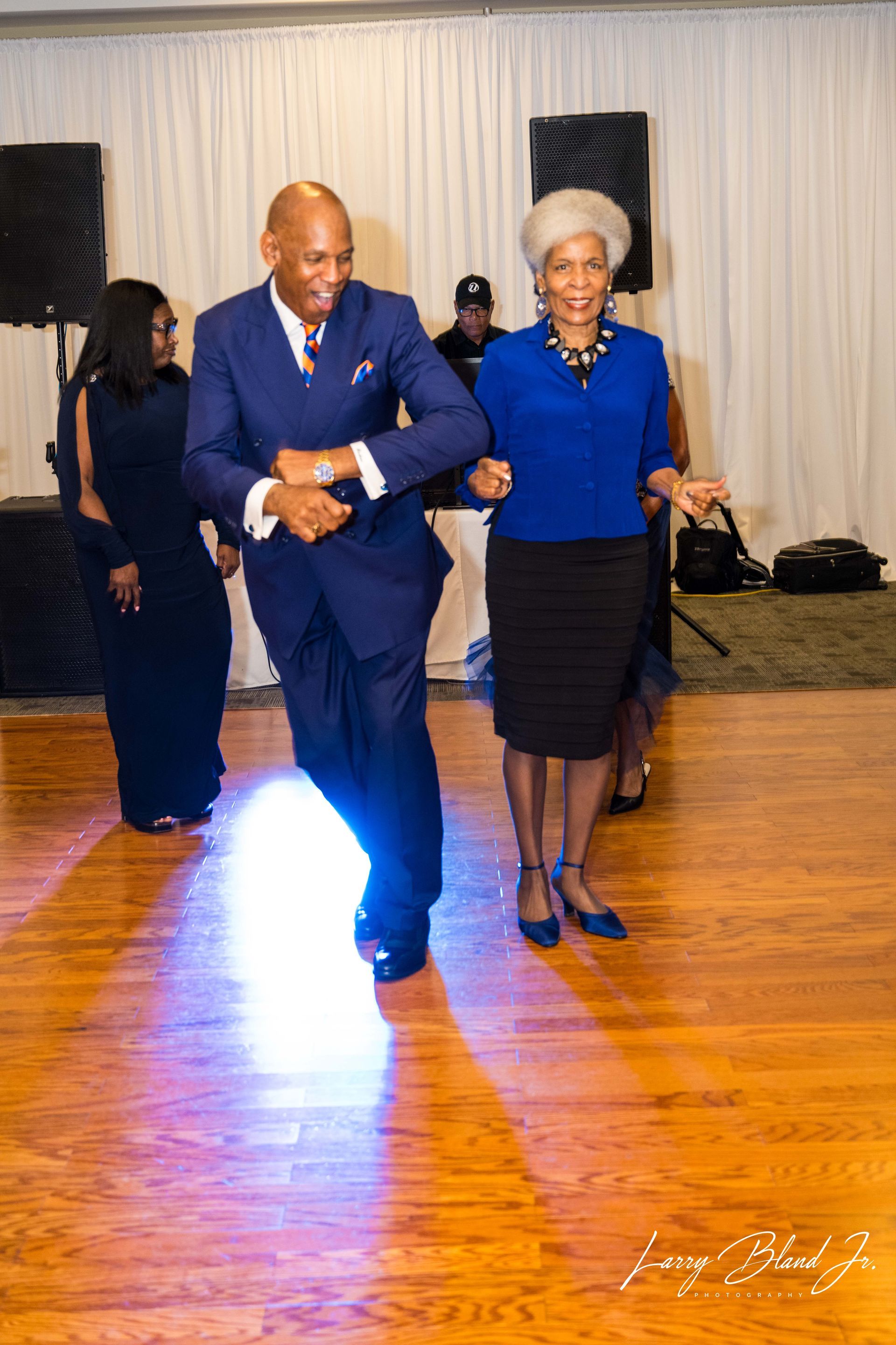 Couple dancing on a wooden floor in a ballroom. Man in blue suit, woman in blue top and black skirt.