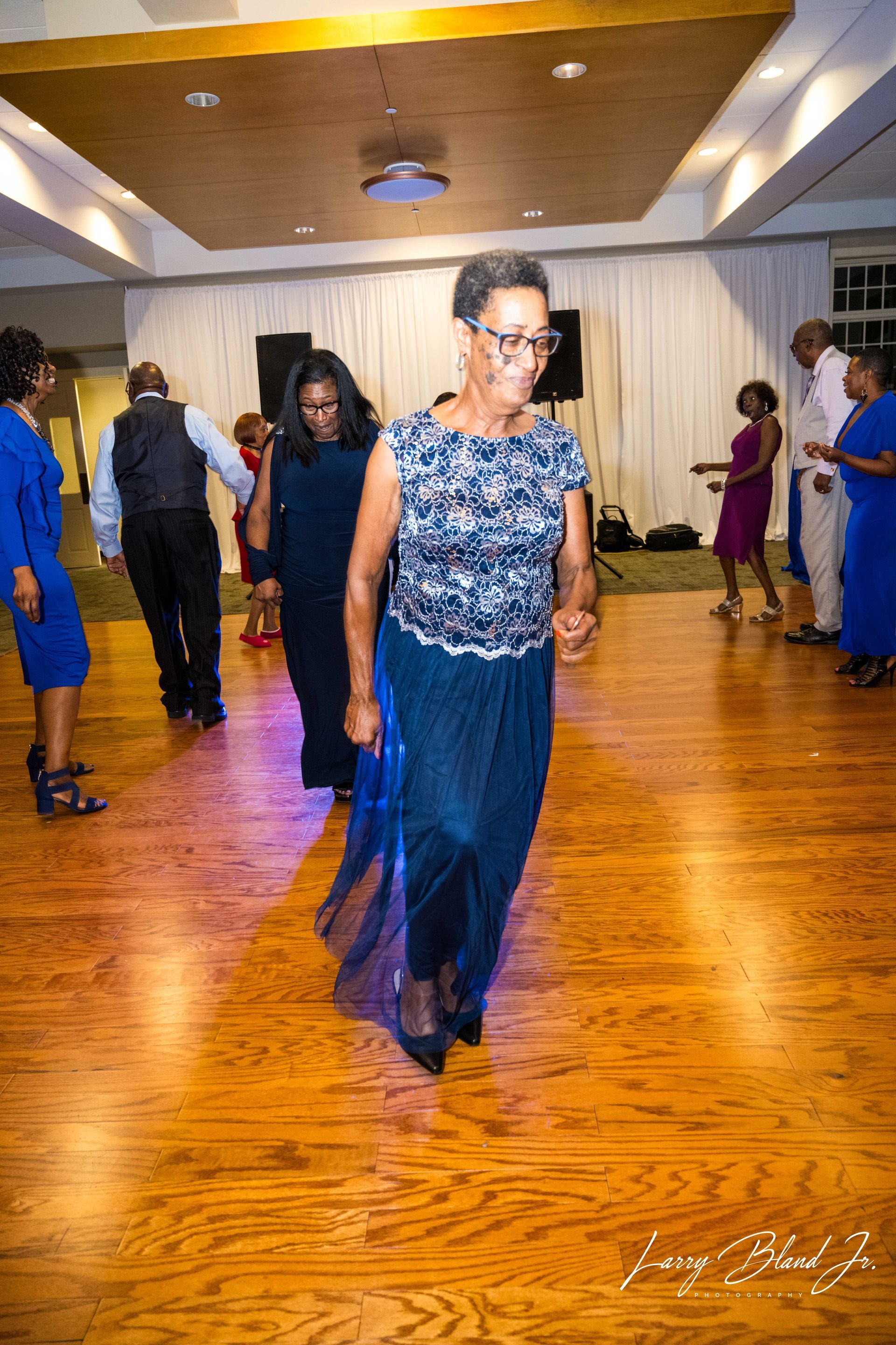 People dancing on a wooden floor in a well-lit room. Woman in blue outfit leading the dance.