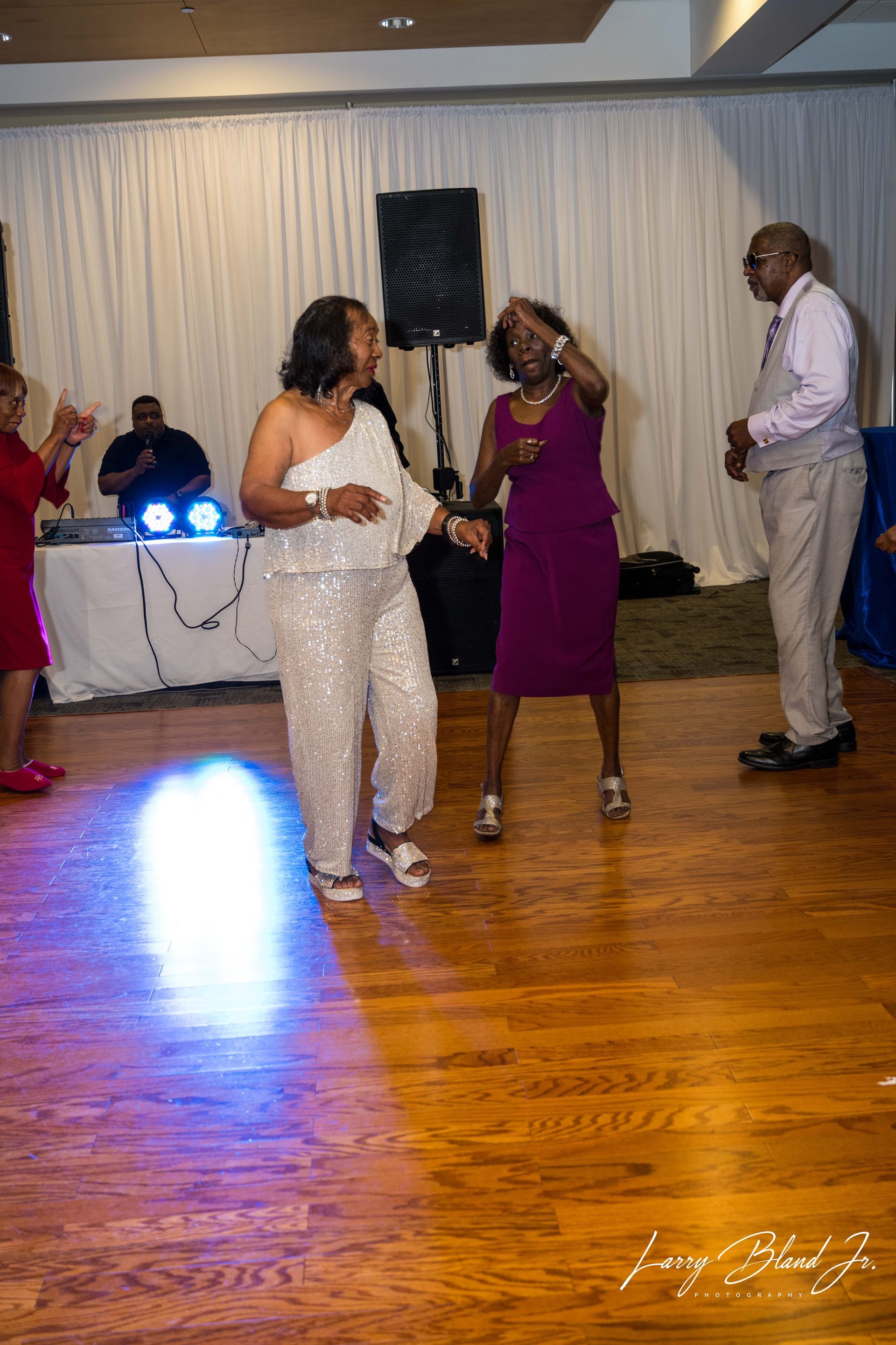 People dancing at a party on a wooden floor, colorful lighting, DJ booth in background.