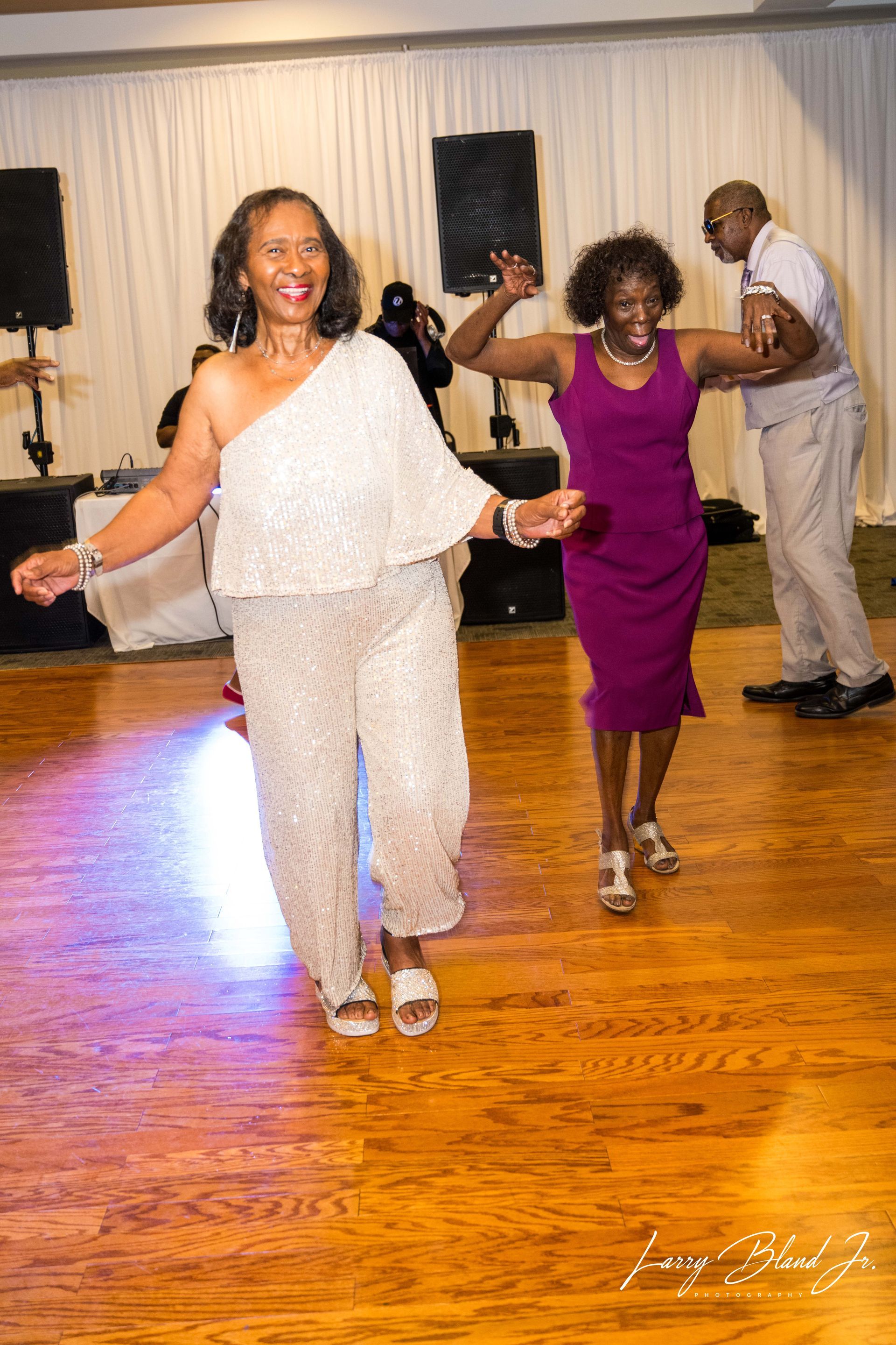 Two women dancing at an event, one in a sparkly jumpsuit, the other in a purple dress, on a wooden dance floor.