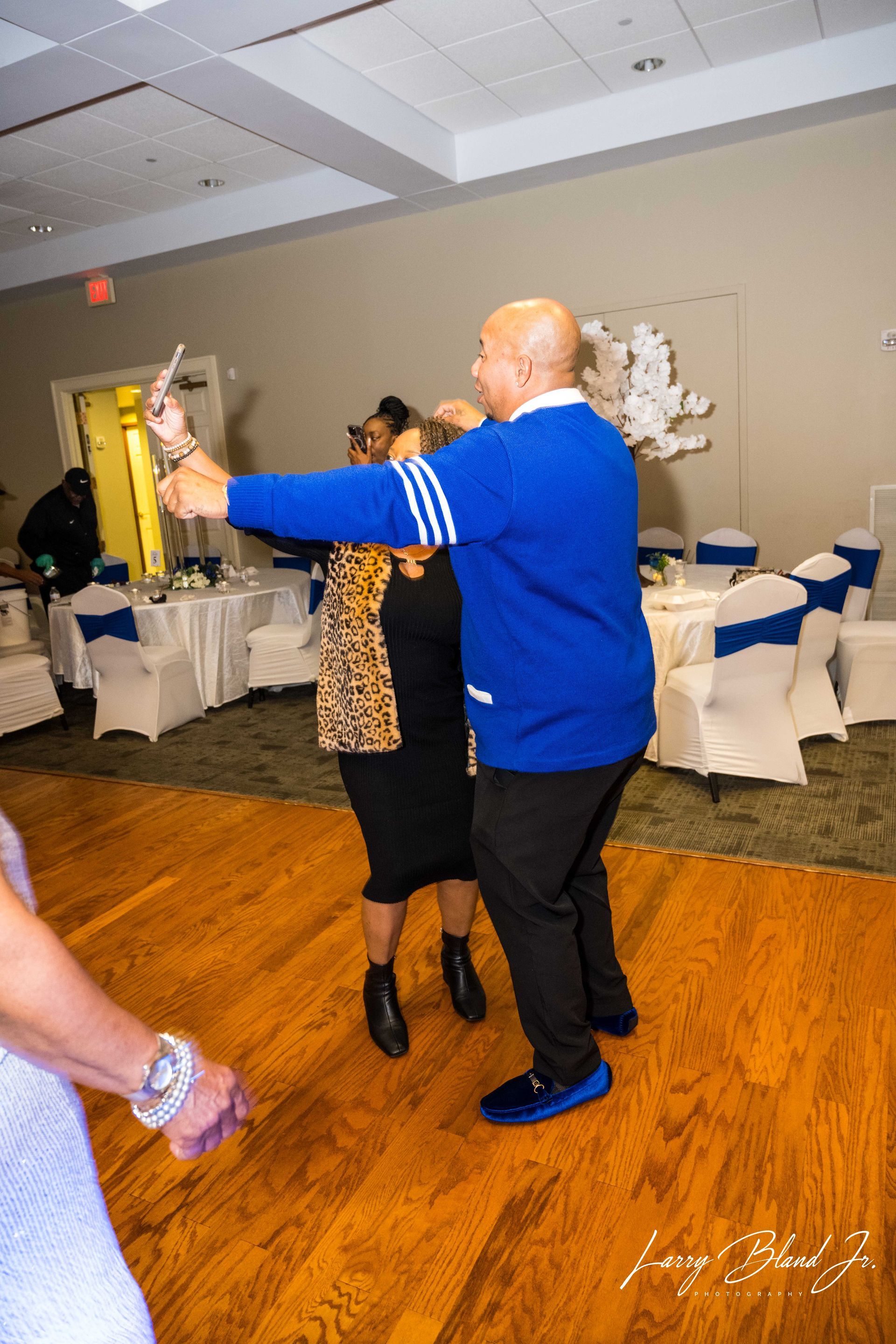 People dancing in a brightly lit ballroom with tables. A man in blue sweater dances with a woman in black dress.