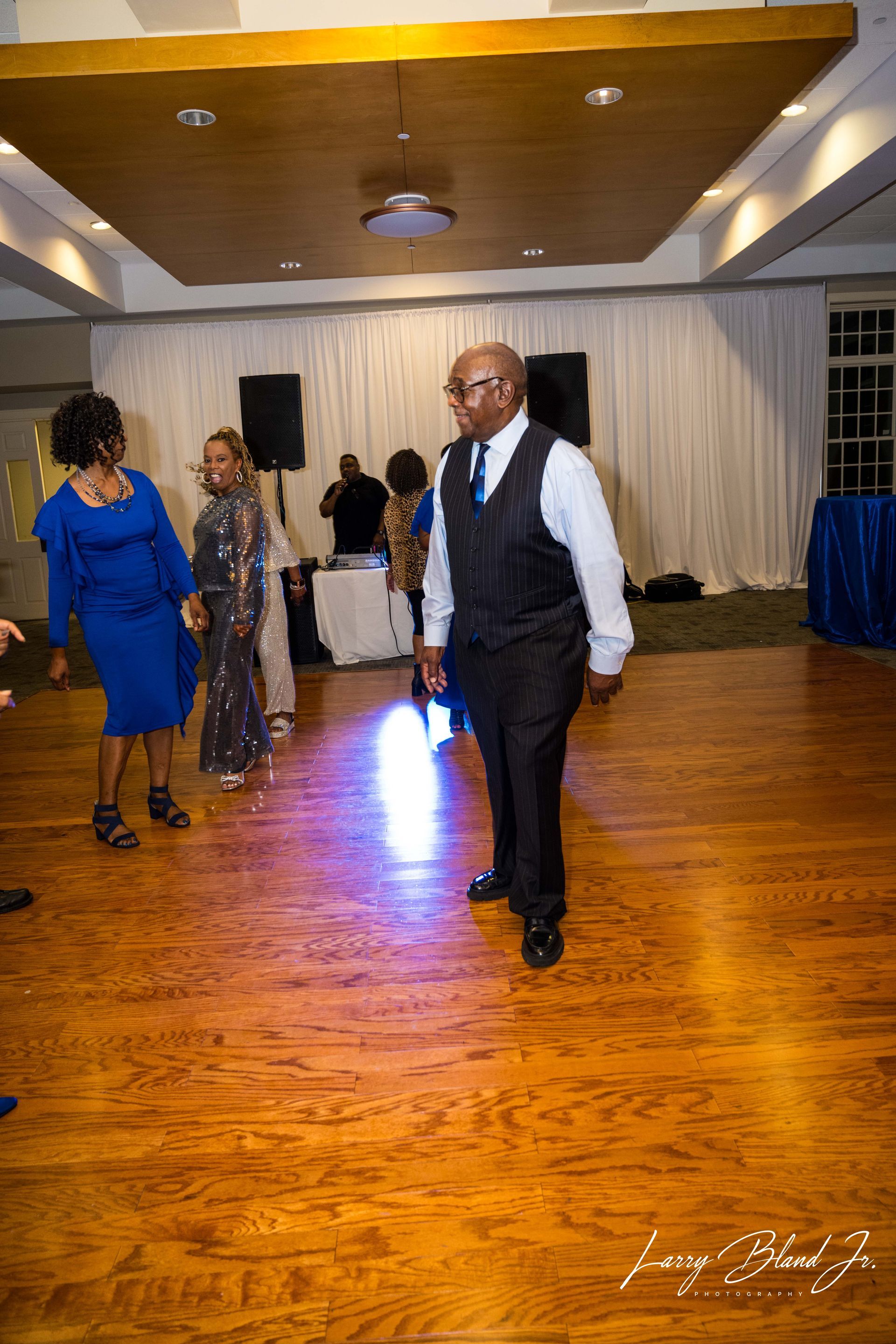 Man in vest dancing at an event with other people in a room with wood flooring.
