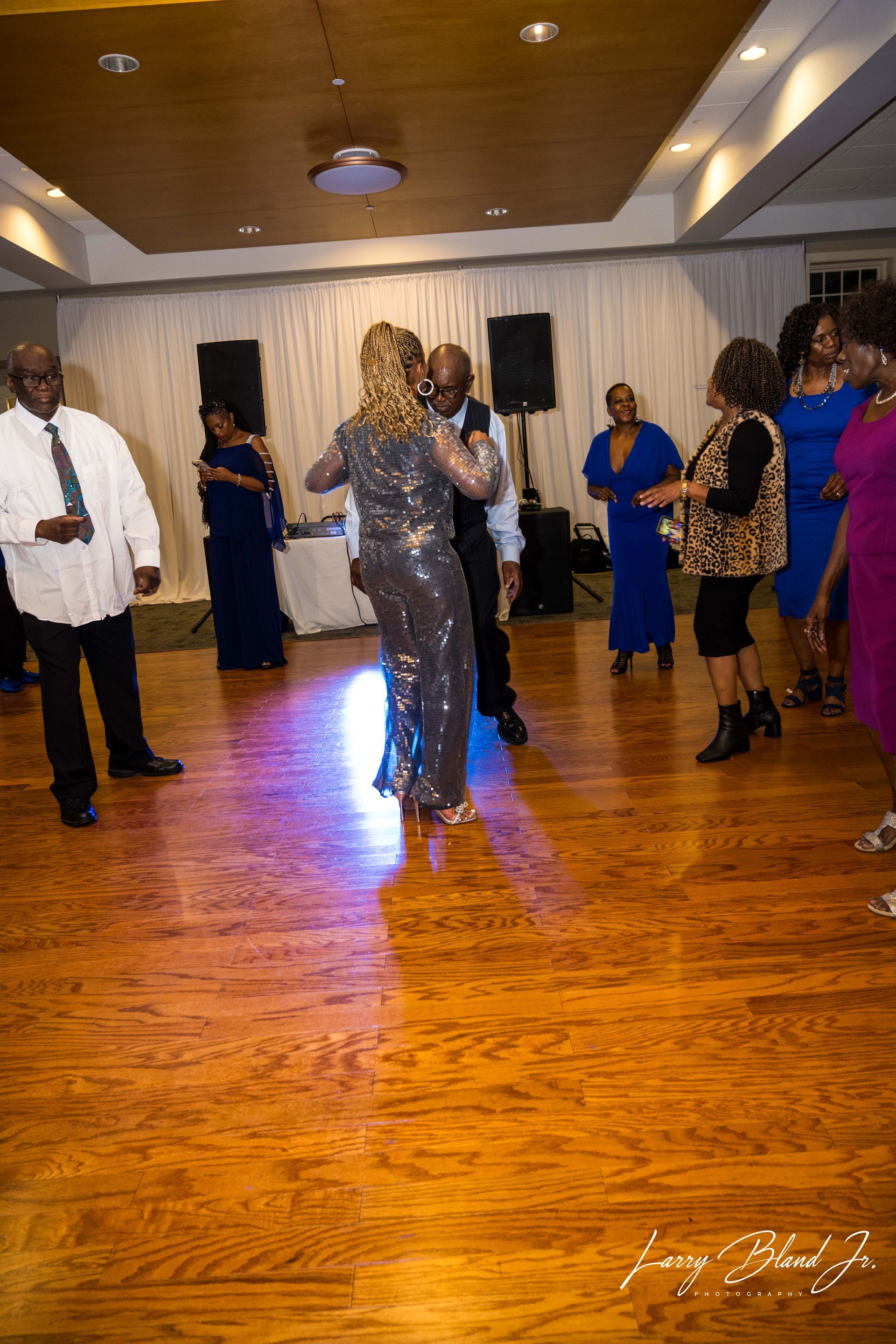 Couple dancing on a wooden dance floor at an event; other people watching nearby.