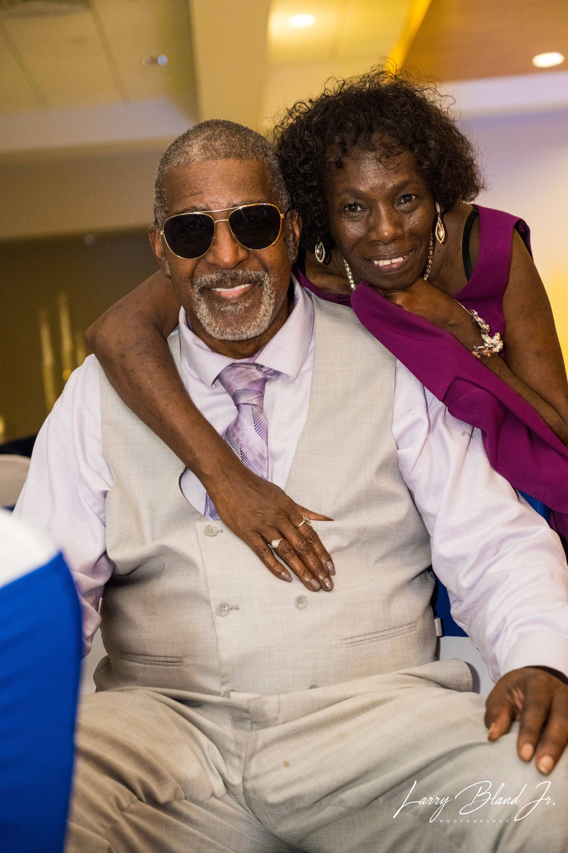 Man in vest and sunglasses smiles, arm around woman in purple, both smiling. Formal setting.