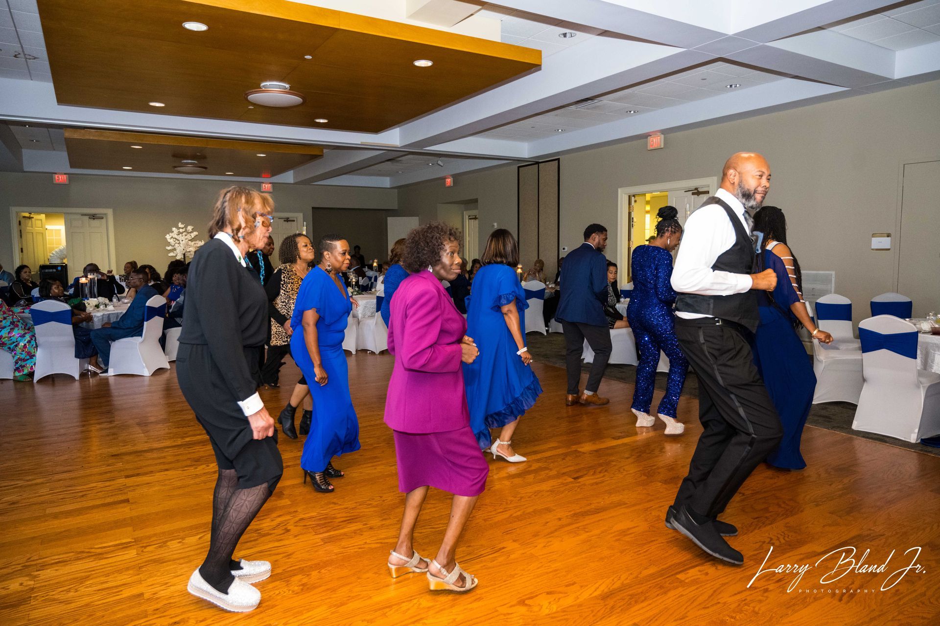 People dancing in a brightly lit ballroom with tables. Some are wearing blue and pink outfits.