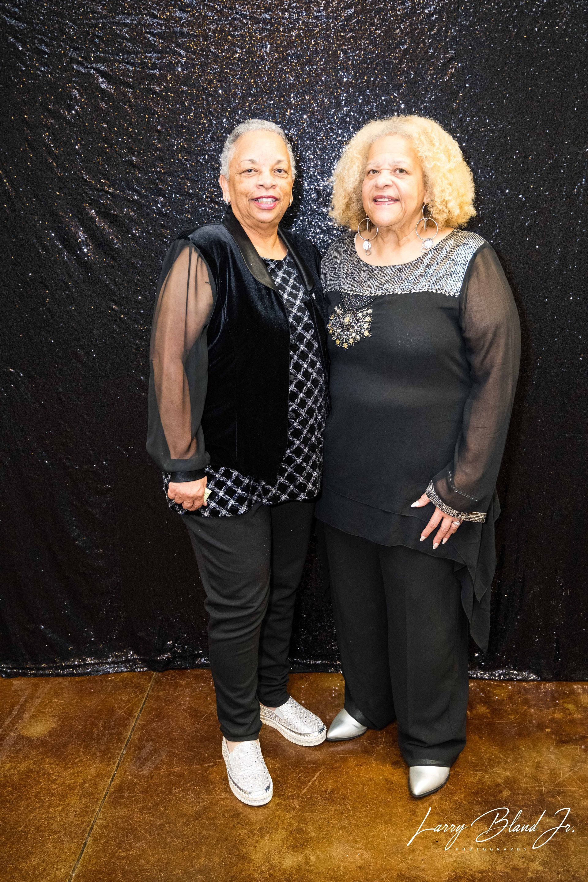 Two women posing in front of a dark, glittery backdrop. One wears black, the other a black vest.