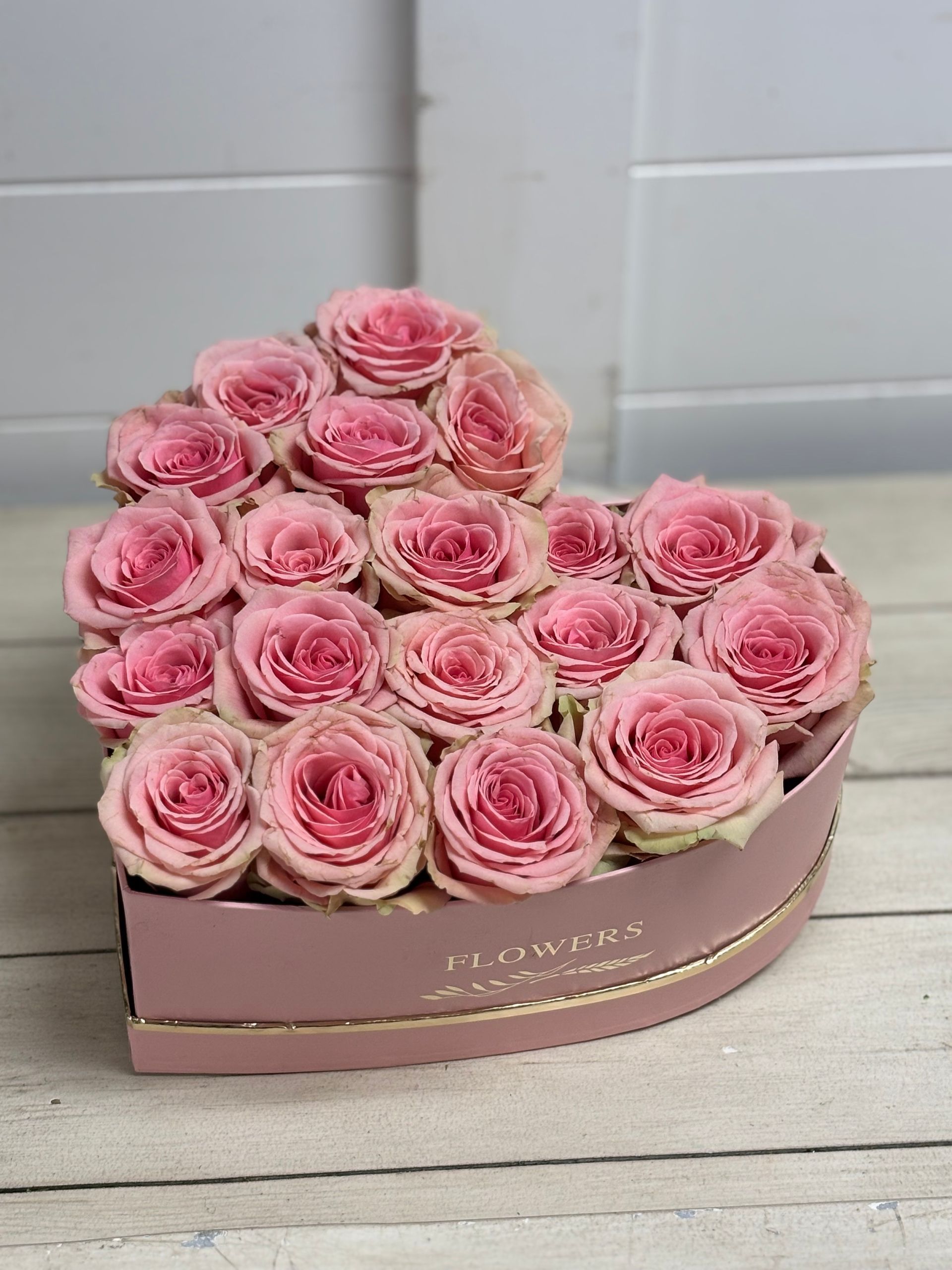 A heart-shaped gift box filled with light pink roses, resting on a white wooden surface.