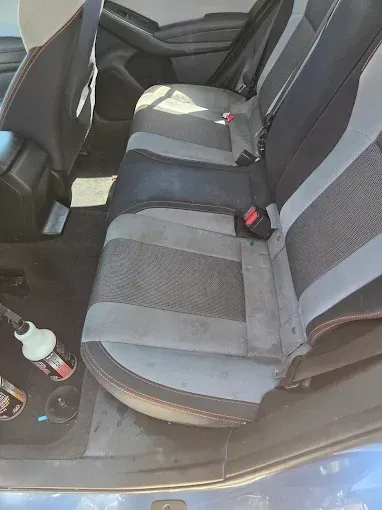Backseat of a blue car with gray and black patterned upholstery and red stitching. Cleaning products sit nearby.
