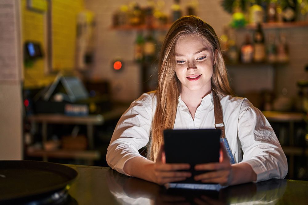 Woman in a white shirt smiling at a tablet in a dimly lit bar, with a blurred red light in the background