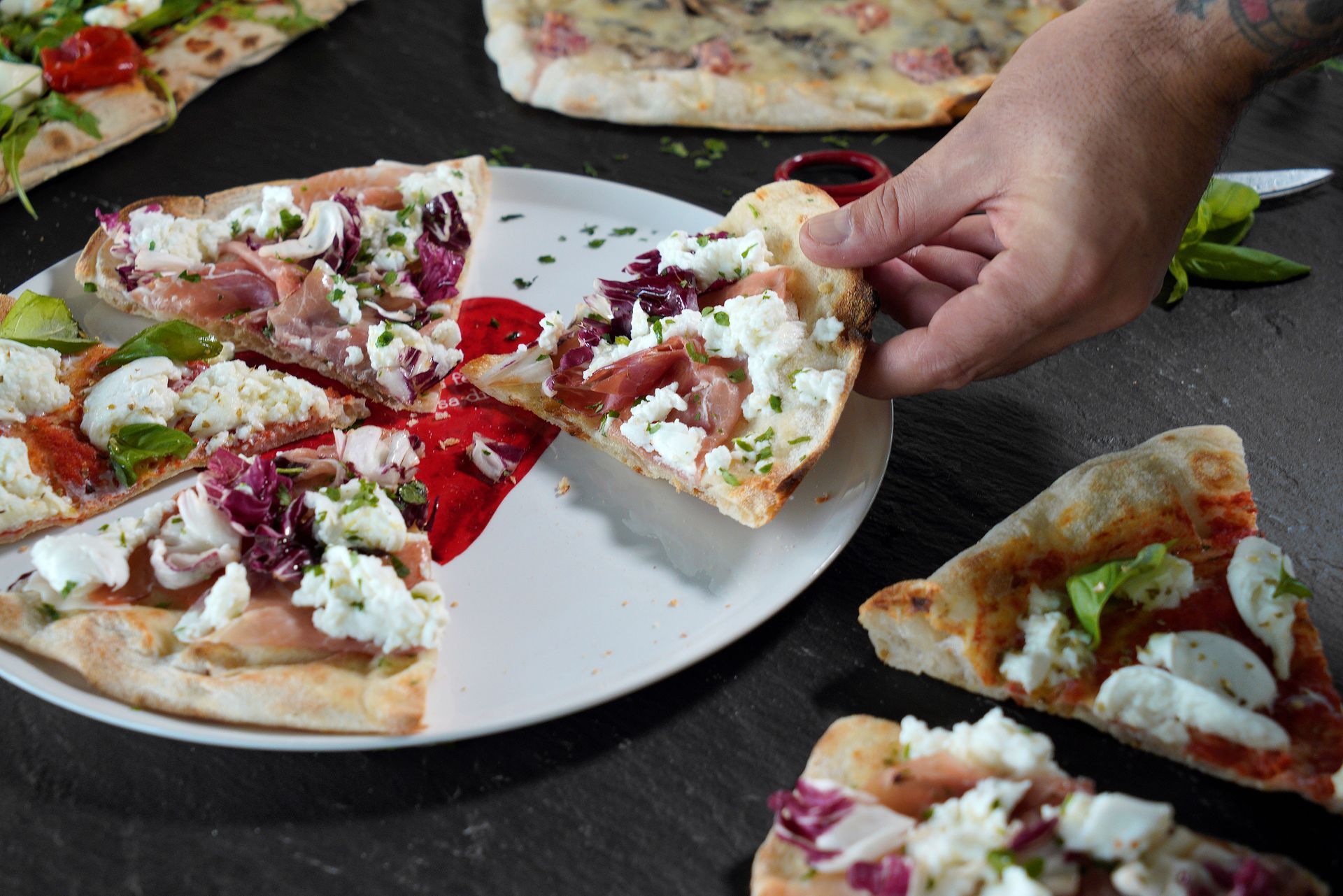 Hand reaching for a slice of veggie pizza on a plate, with more slices on a dark countertop.