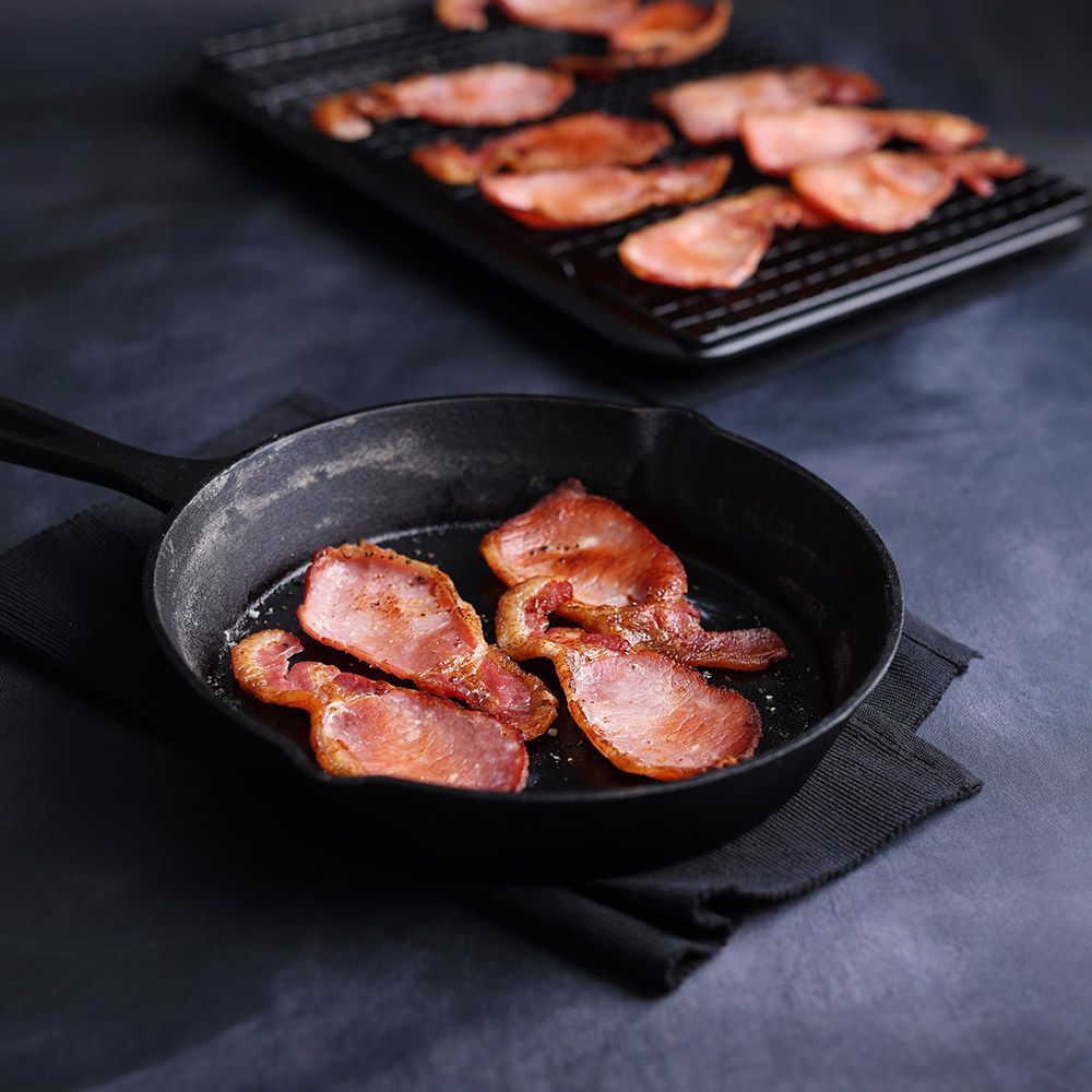 Bacon strips sizzling in a skillet, with more bacon cooking on a rack in the background.