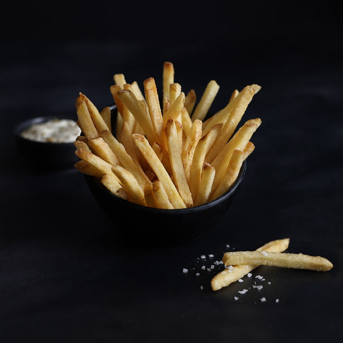 Bowl of golden French fries on a dark background, with a few fries and salt scattered nearby