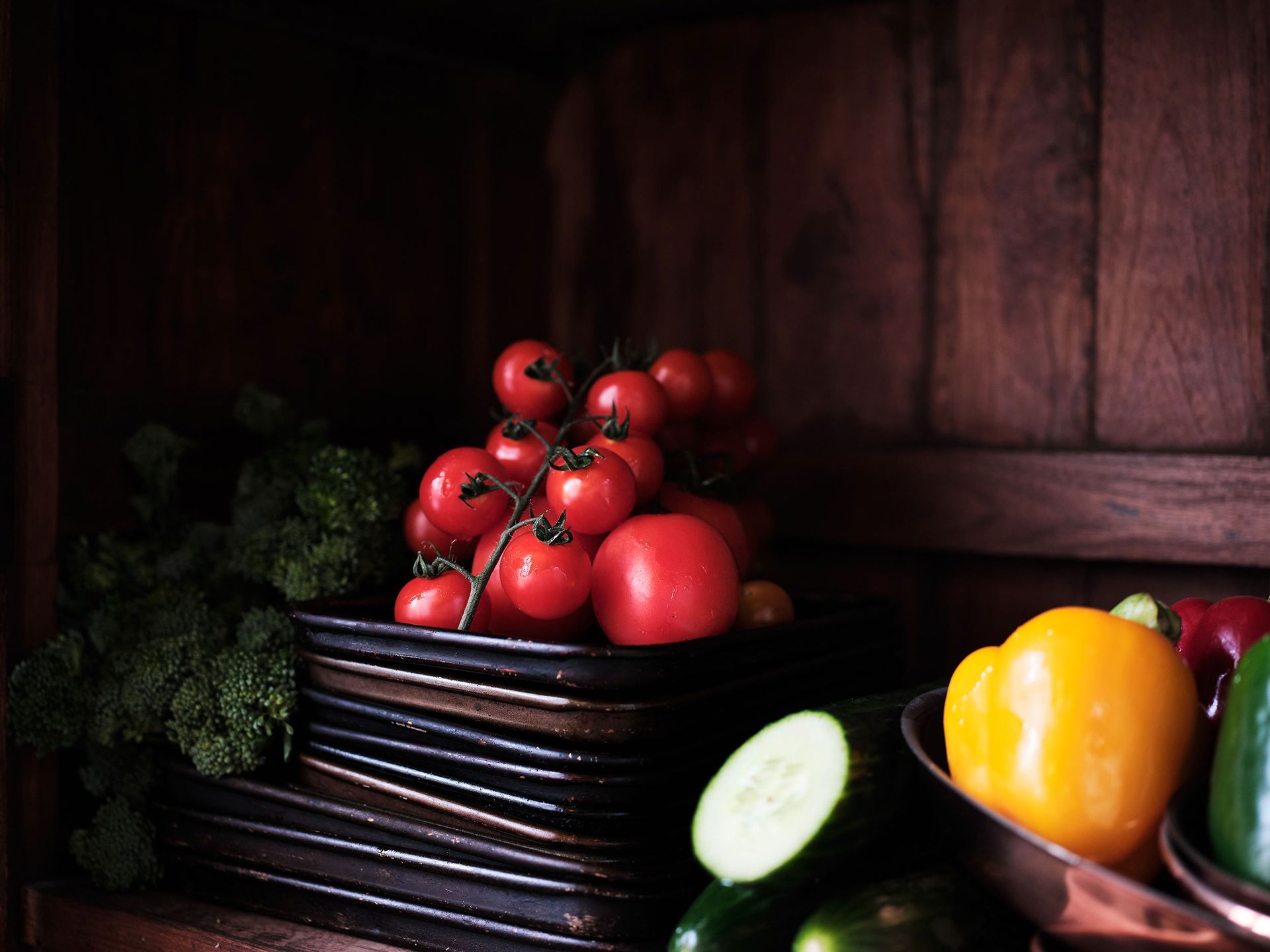 Basket of tomatoes, cucumbers, and peppers on a dark wooden table in a rustic kitchen