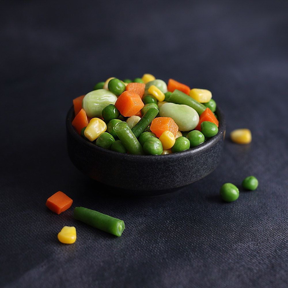 Black bowl of mixed frozen vegetables on a dark surface, with a few pieces scattered around it