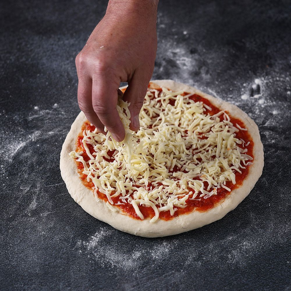 Hand sprinkling shredded cheese onto a pizza with tomato sauce on a floured dark surface