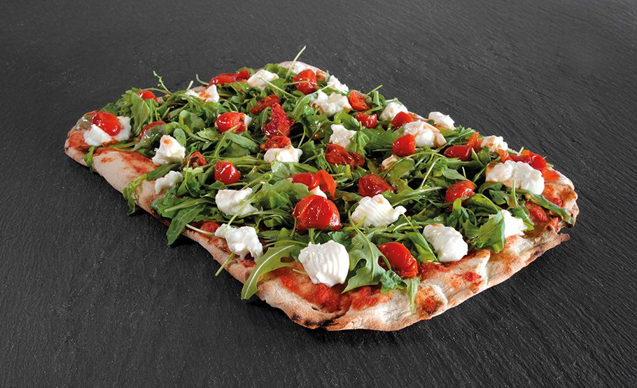 Rectangular flatbread pizza topped with greens, cherry tomatoes, cheese, and white sauce on a dark surface