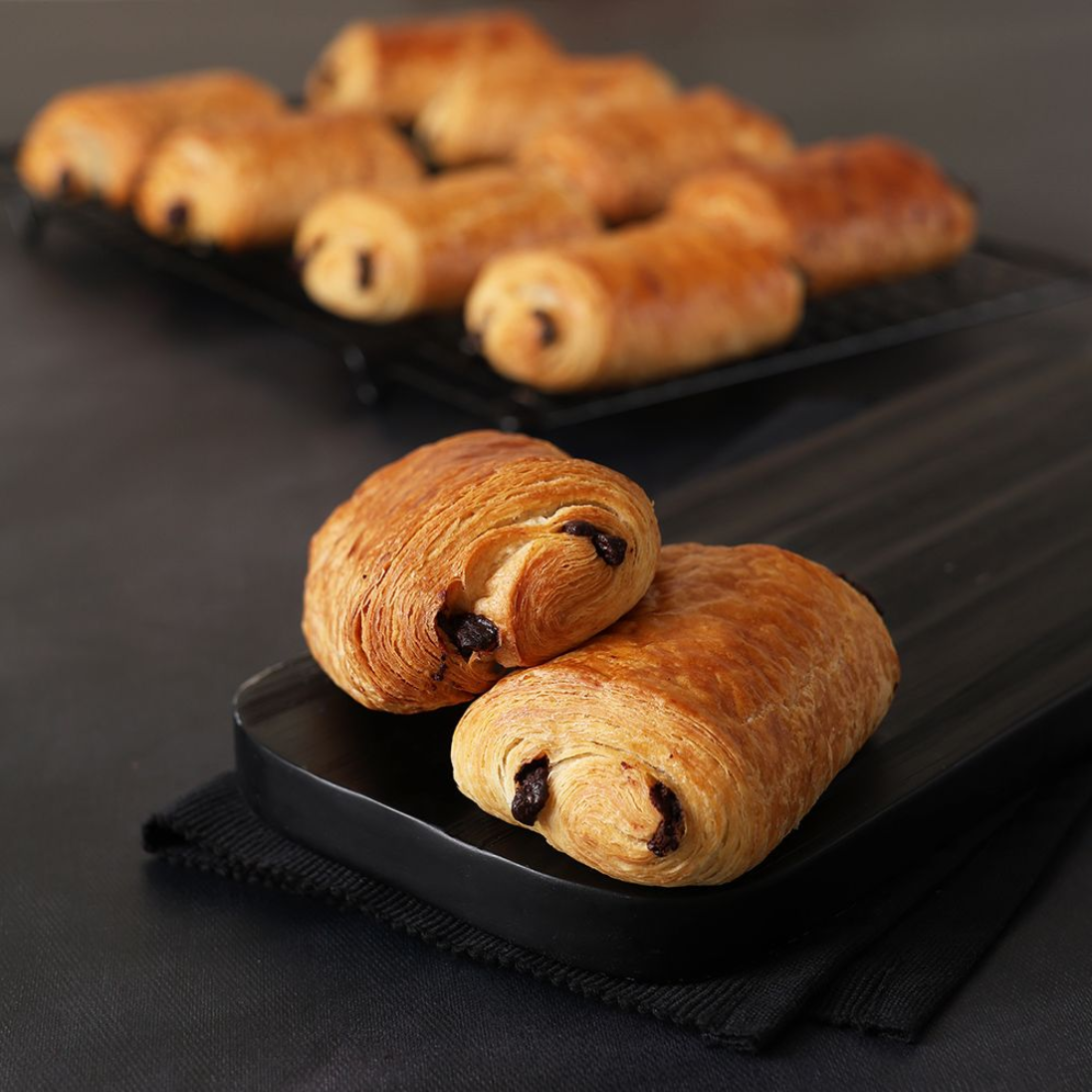 Chocolate croissants on a black tray, with more pastries blurred in the background