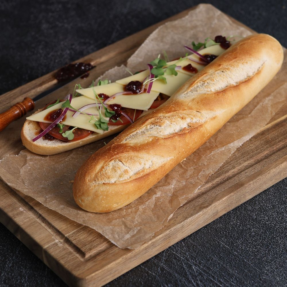 Long baguette sandwich on parchment on a wooden board, topped with sliced cheese, red onion, and greens