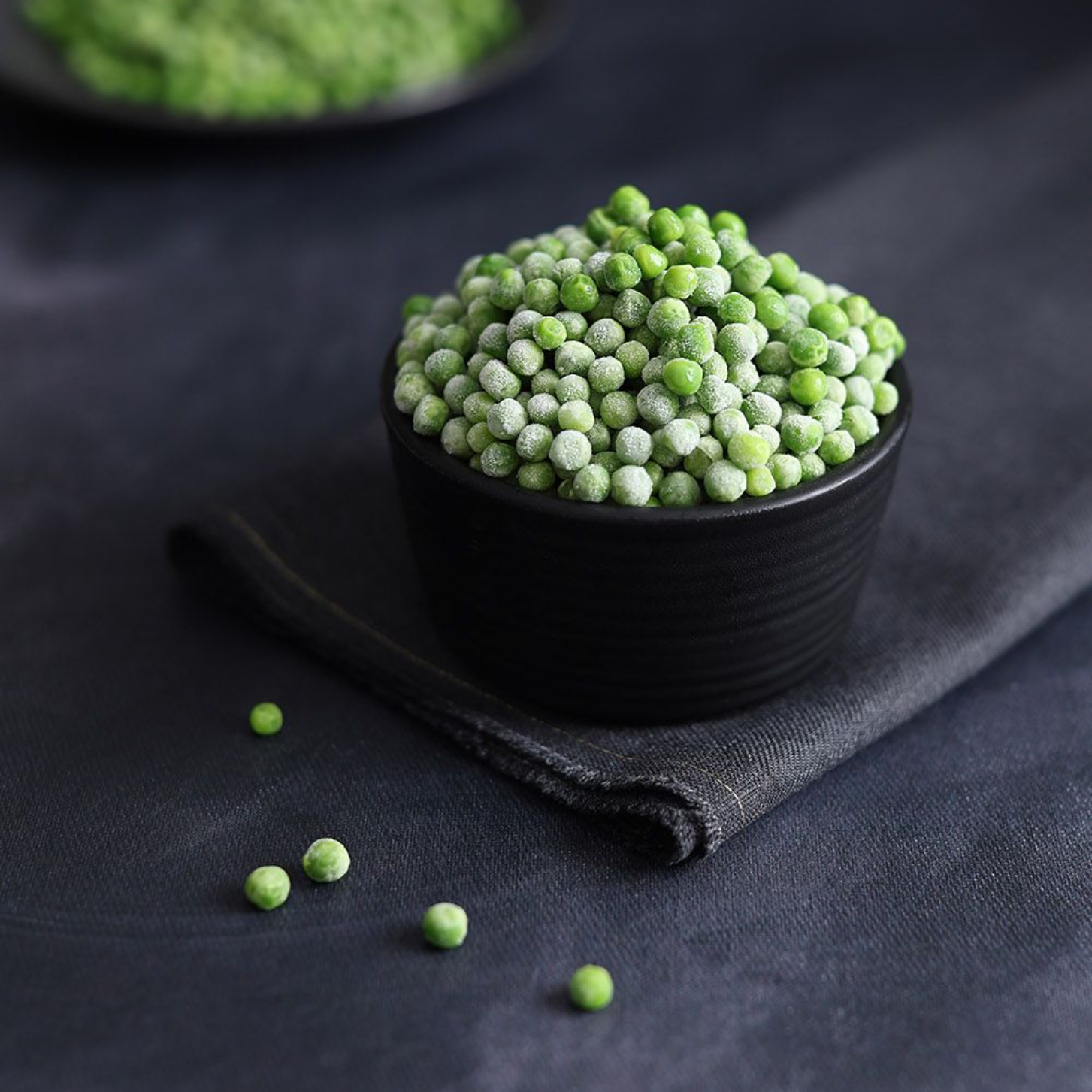 Black cup filled with green peas on dark fabric, with a few peas scattered nearby