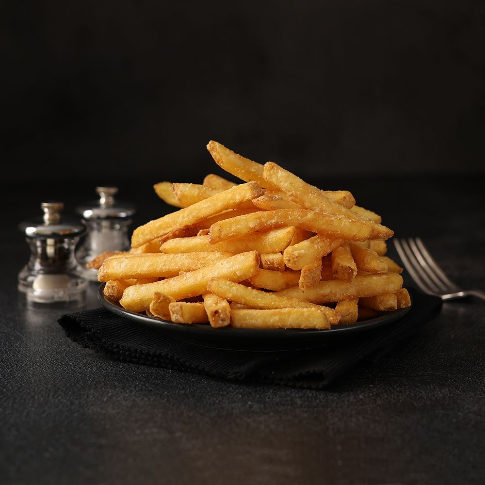 Plate of golden French fries on a dark table with salt shakers and a fork in the background