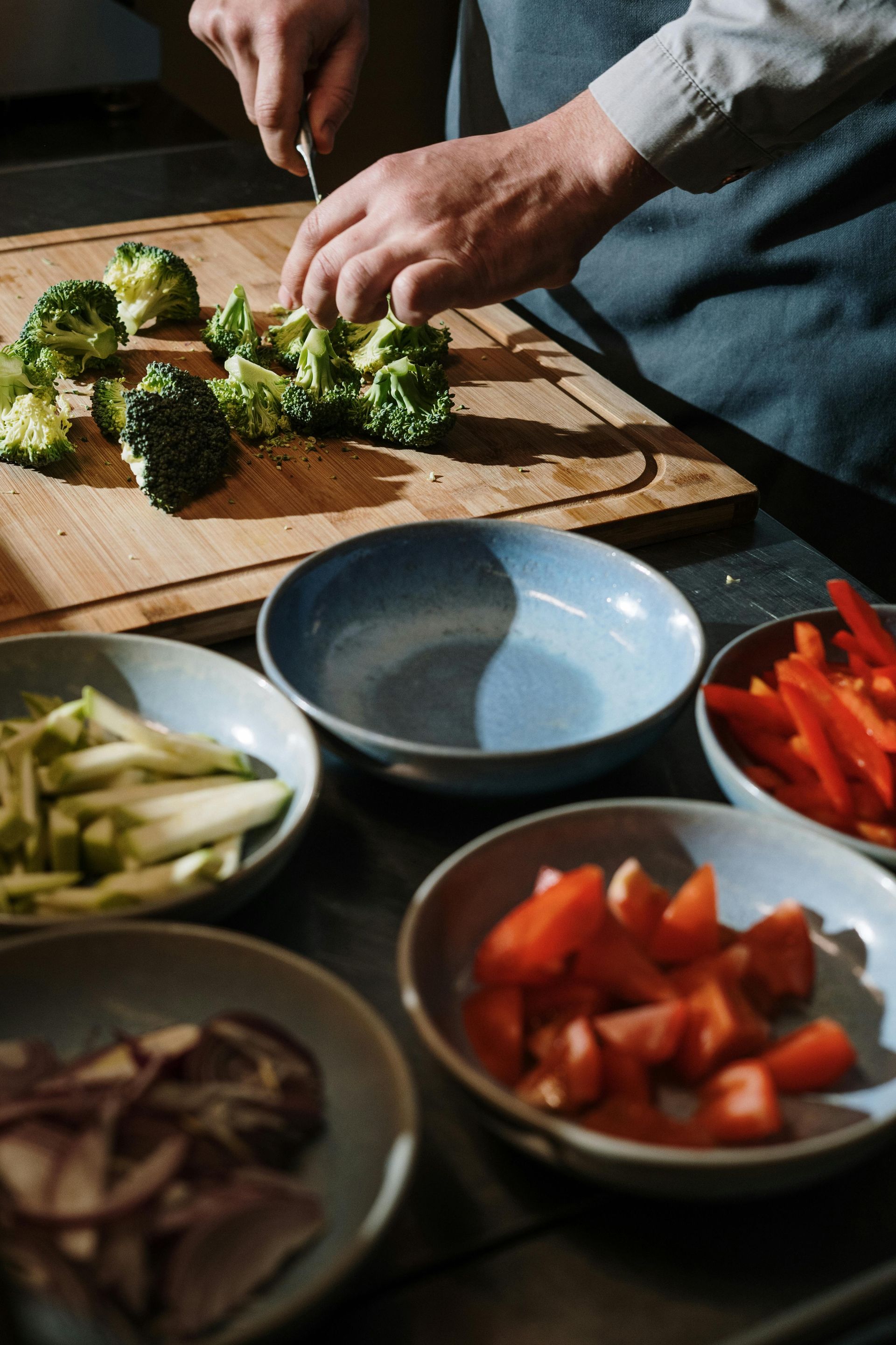 Hands chopping broccoli on a wooden cutting board with bowls of sliced vegetables nearby