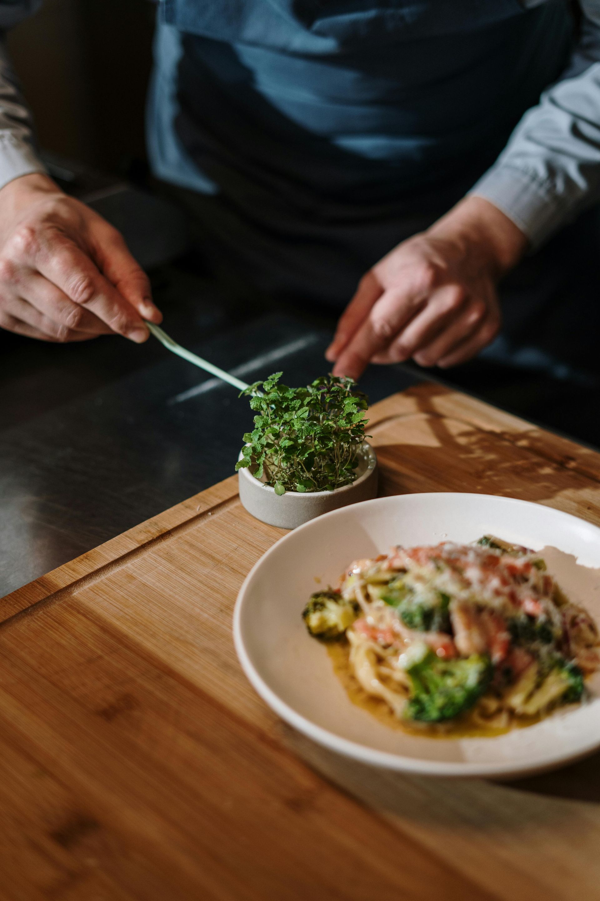 Person plating a gourmet dish beside a small potted herb on a wooden table