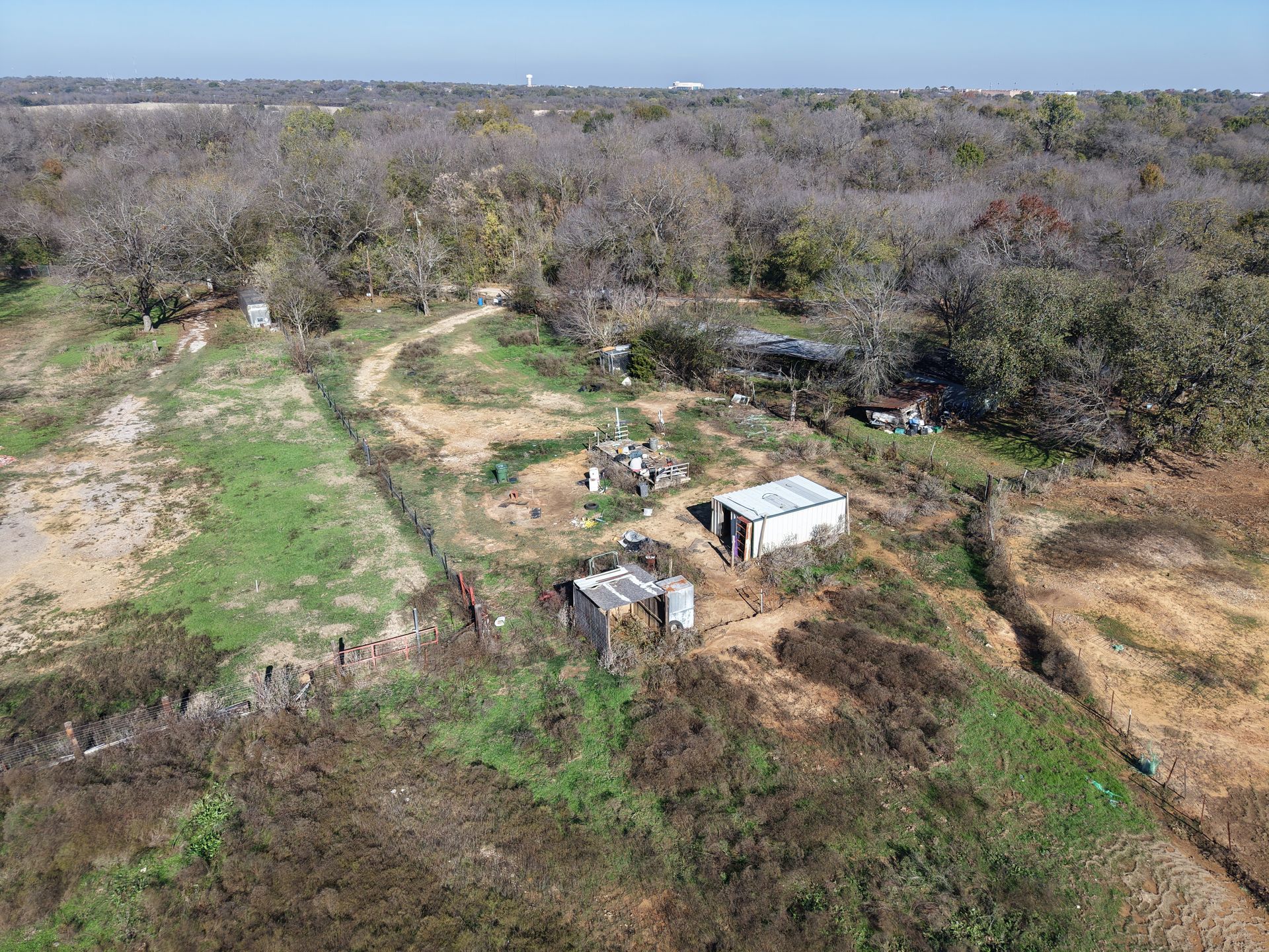 Aerial view of a rural clearing with small white structures, dirt paths, and dense trees.