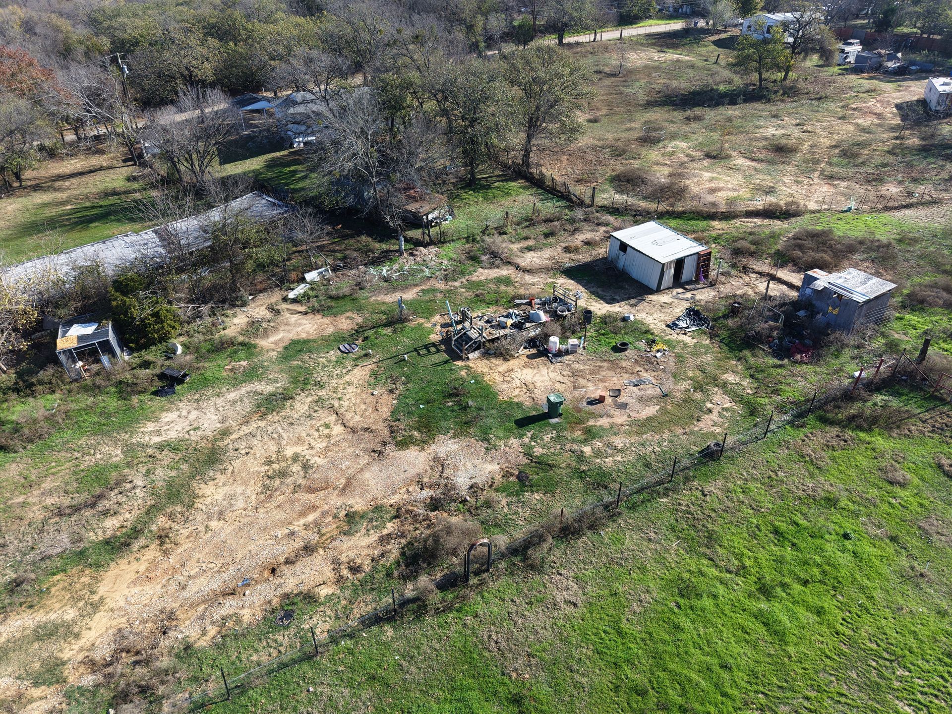Aerial view of a rural yard with a small shed, scattered debris, bare trees, and green grass patches