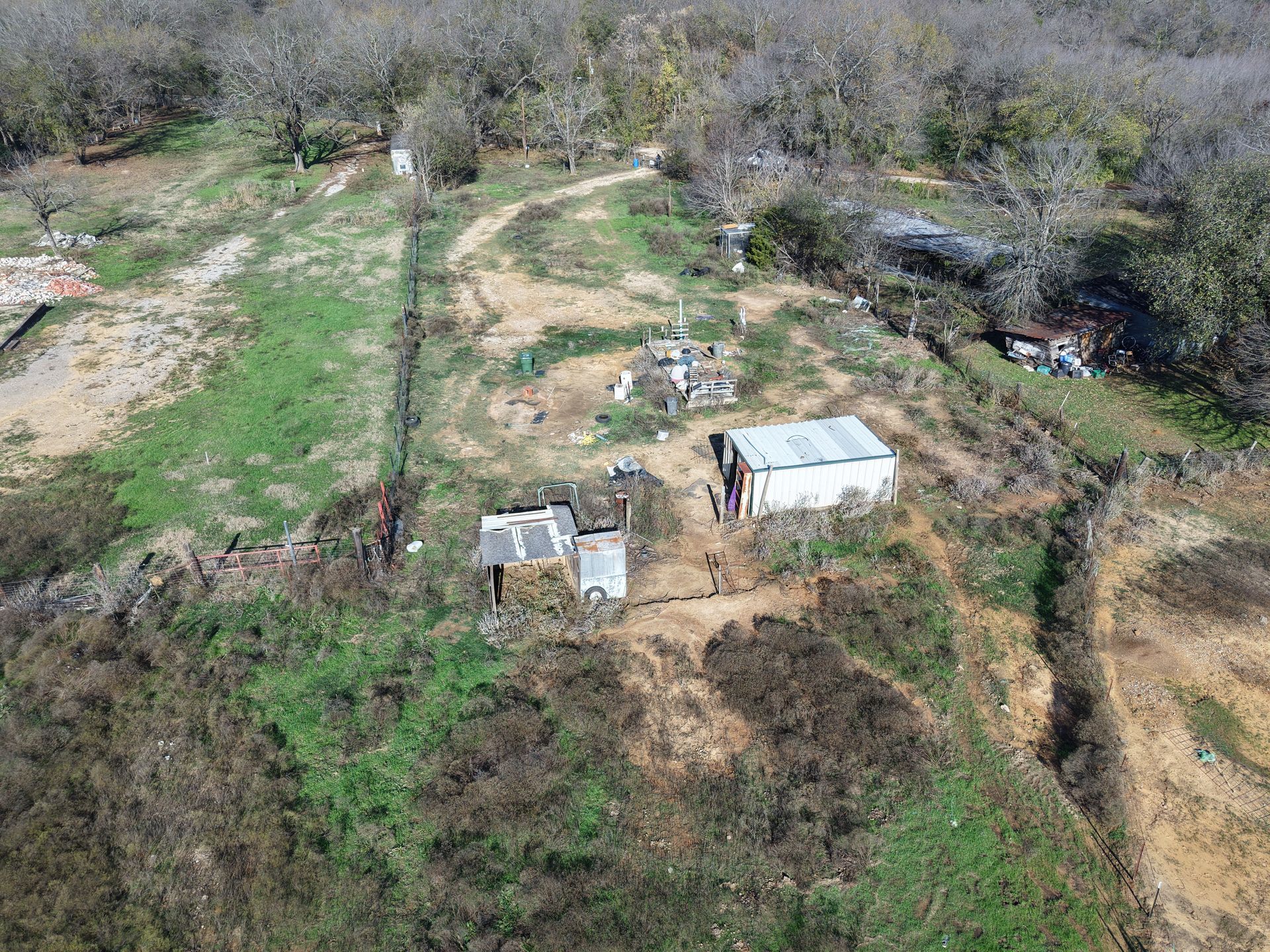 Aerial view of a small rural property with two sheds, dirt paths, and surrounding trees and brush.