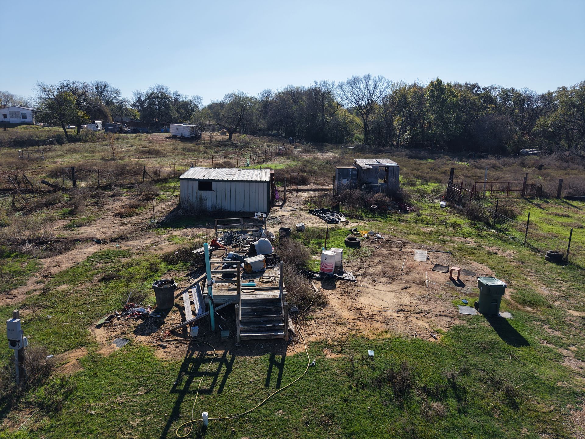 Rural yard with a small white shed, scattered equipment, and bare trees on a sunny hillside