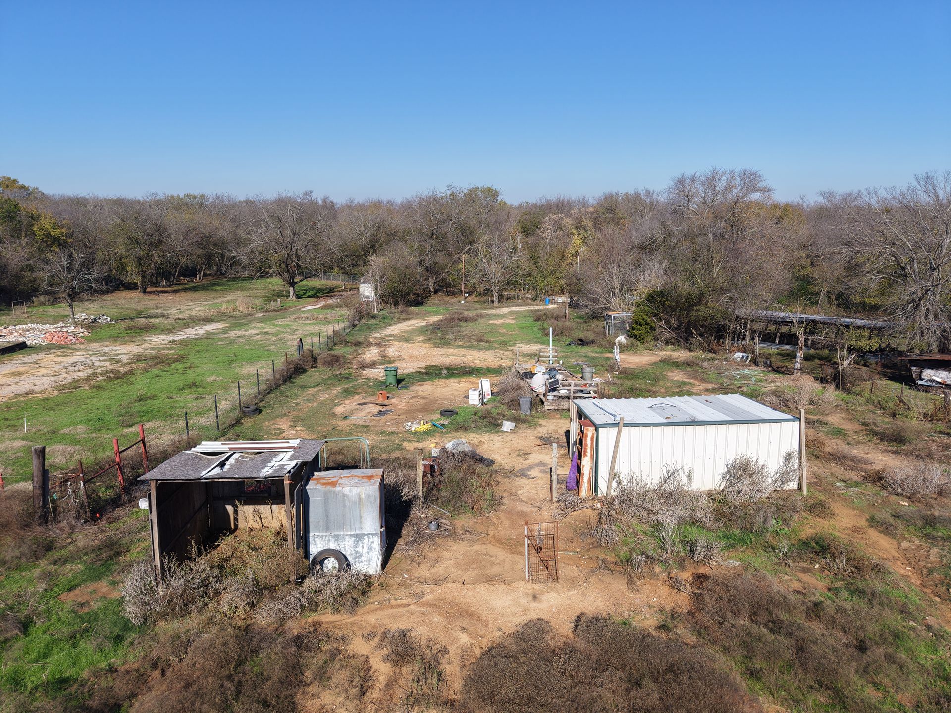 Rural yard with small sheds and a white trailer amid dry grass and trees under a clear blue sky