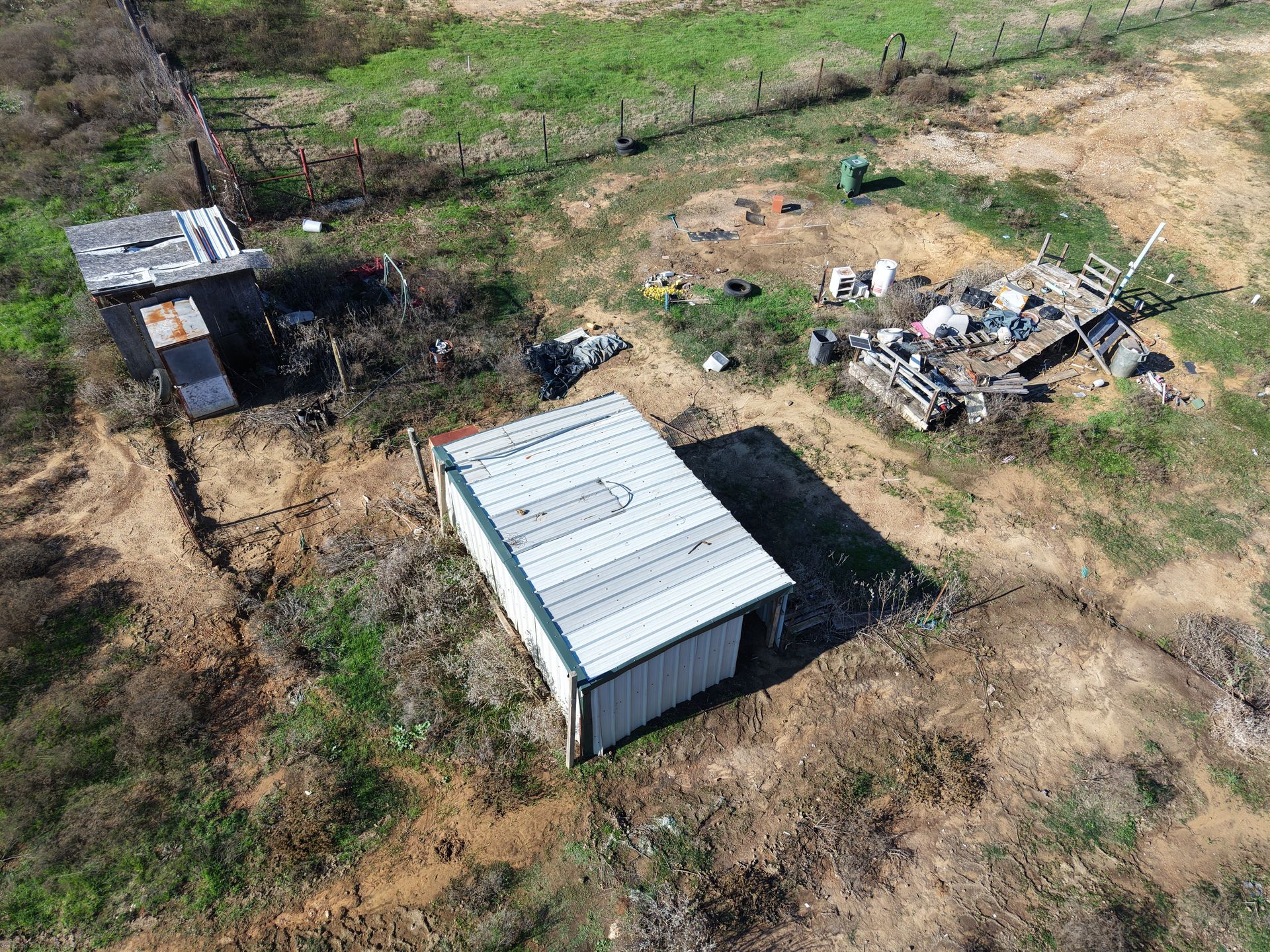Aerial view of a rural site with a small white-roofed shed and scattered debris on dirt and grass.