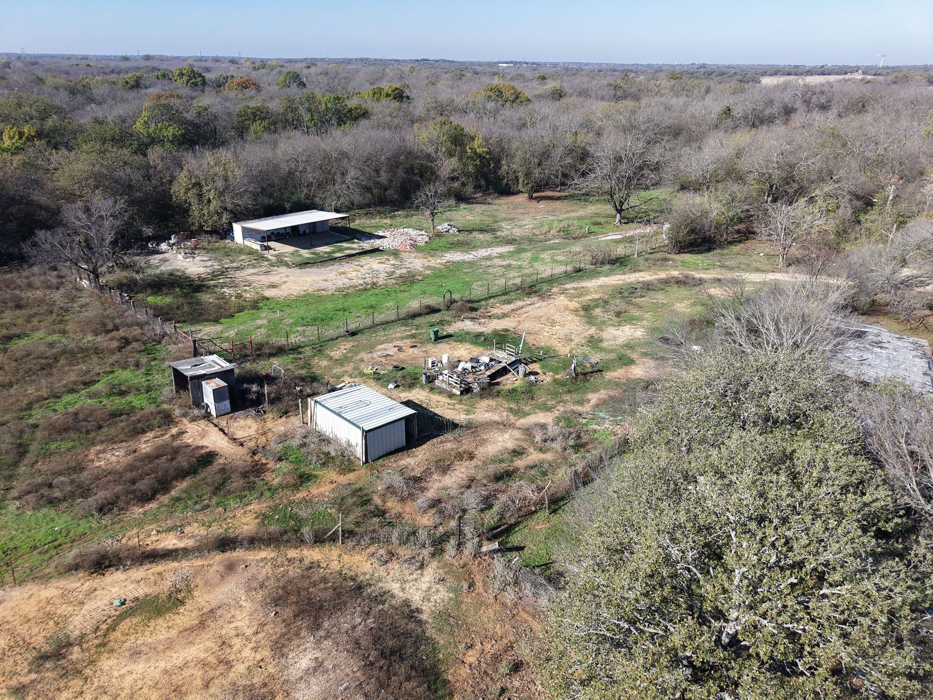 Rural clearing with small sheds and a dirt path surrounded by dry brush and trees.