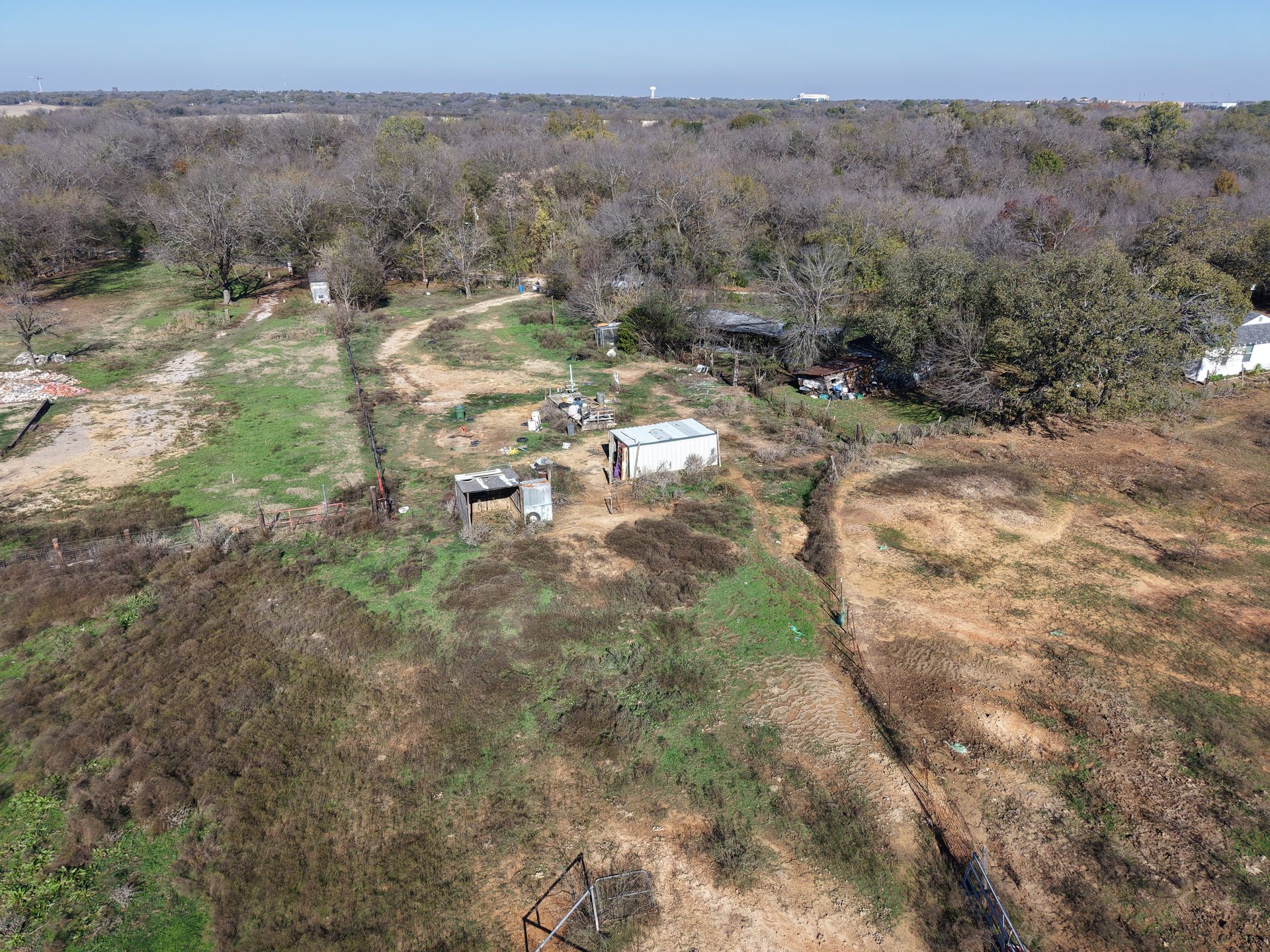 Rural clearing with dirt paths, scattered trees, and a few small structures under a clear blue sky