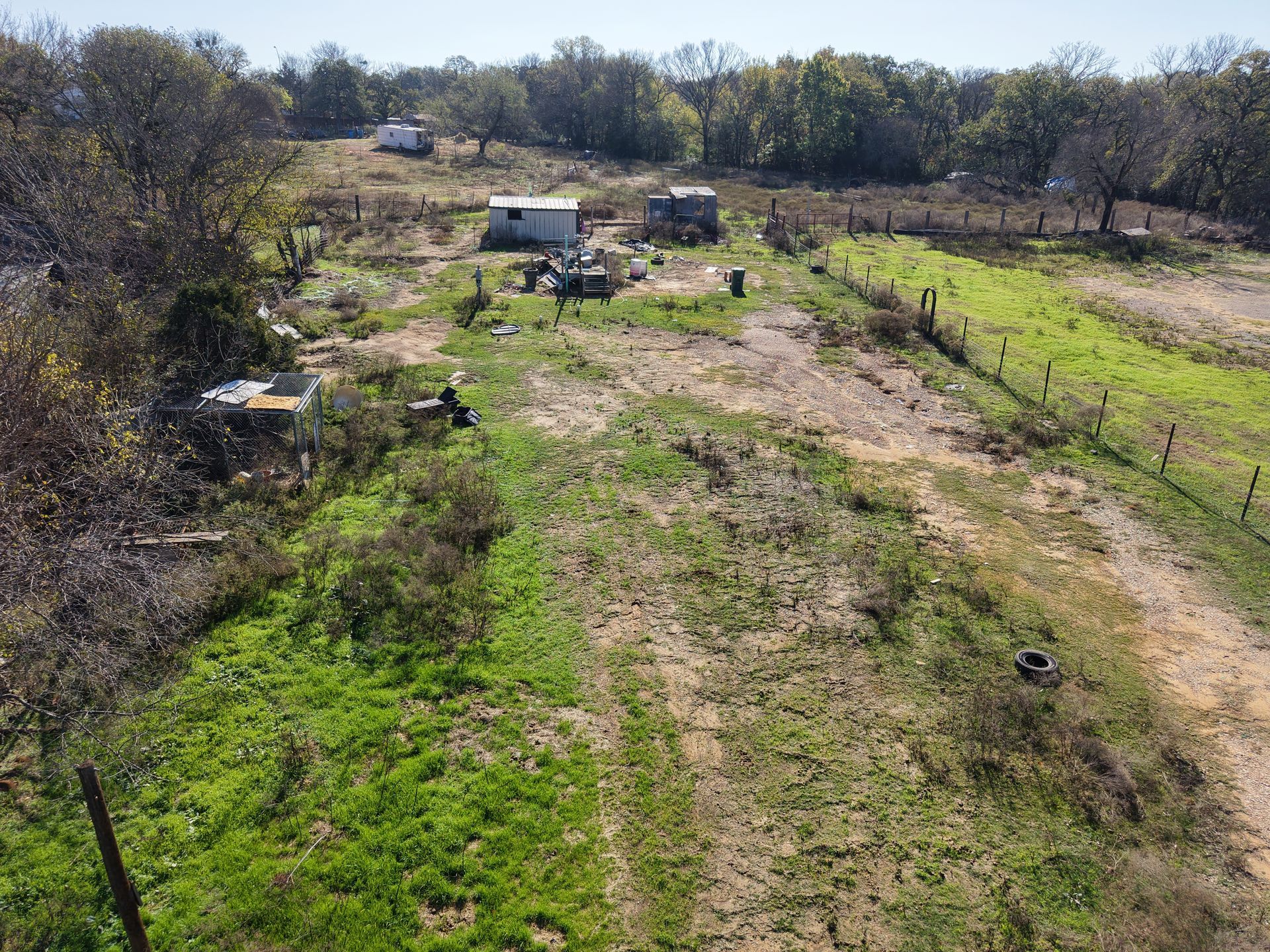 Rural dirt road with a small white vehicle, fences, and scattered trees under a clear sky