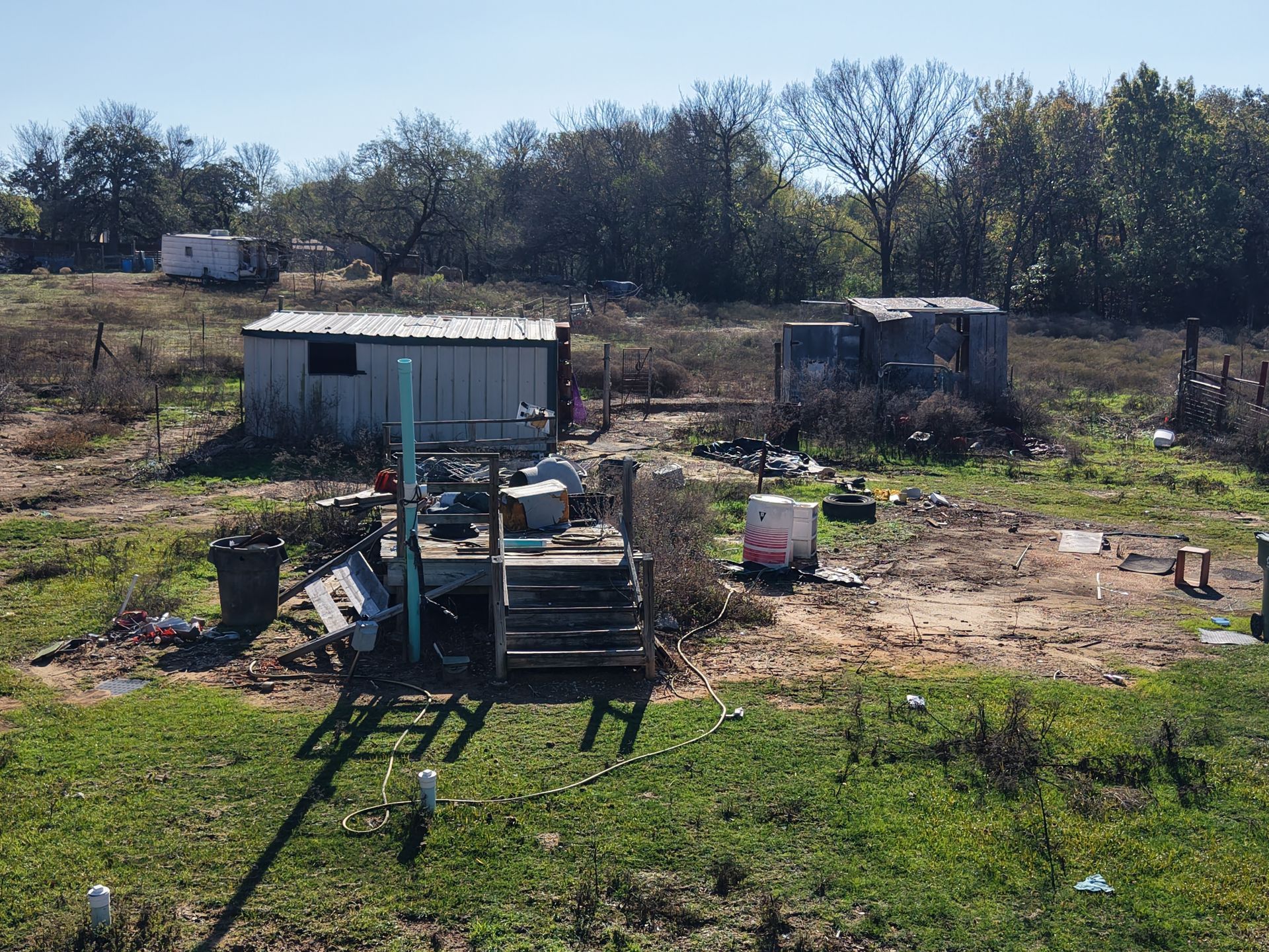 Rural yard with a small shed, scattered tools, and a weathered trailer under trees on a sunny day