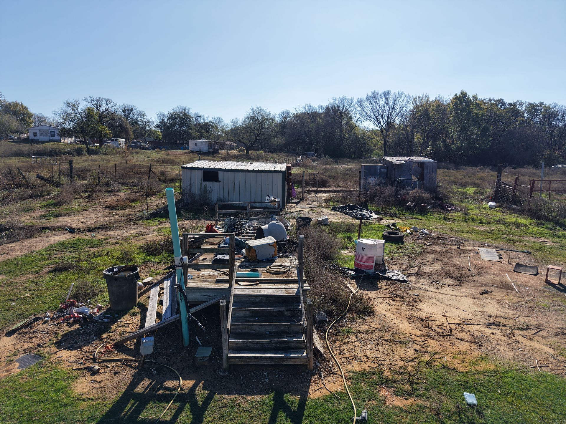 Rural yard with a small wooden platform, scattered debris, and a white shed under a clear sky.