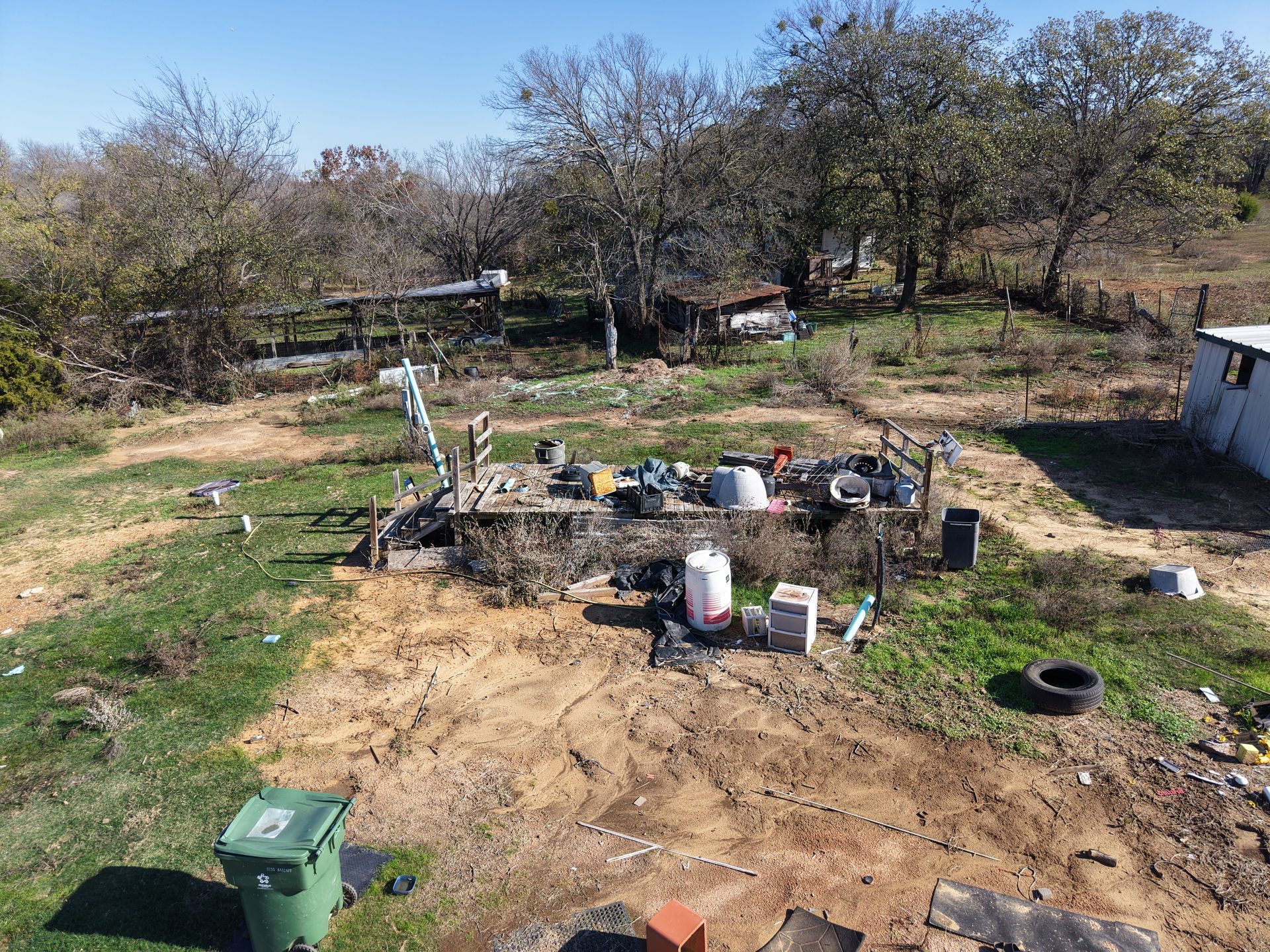 Small outdoor worksite with scattered equipment, buckets, tires, and a green utility box in a dry, brushy yard.