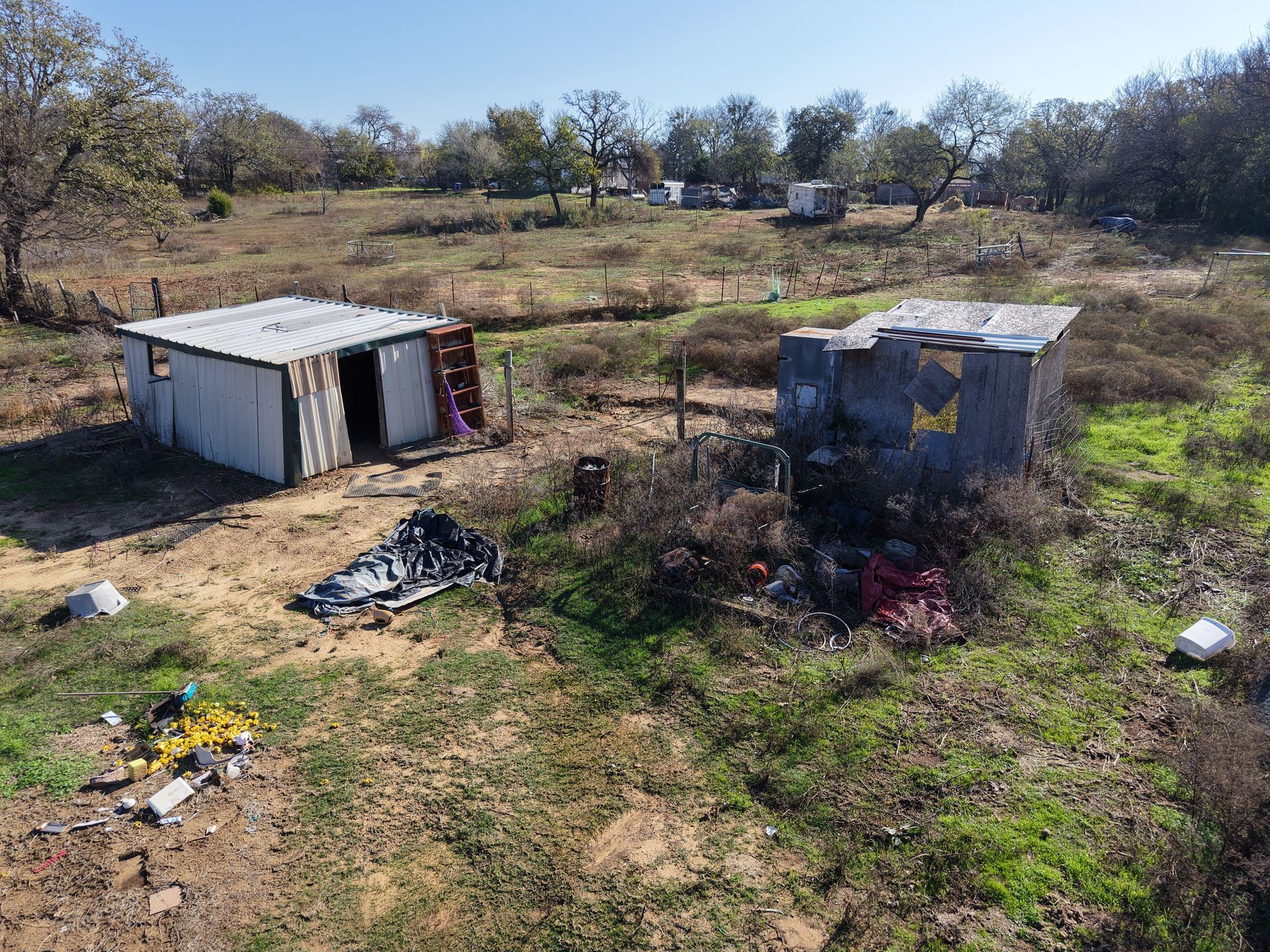 Two small weathered sheds in a dry, grassy field with scattered debris and trees in the background