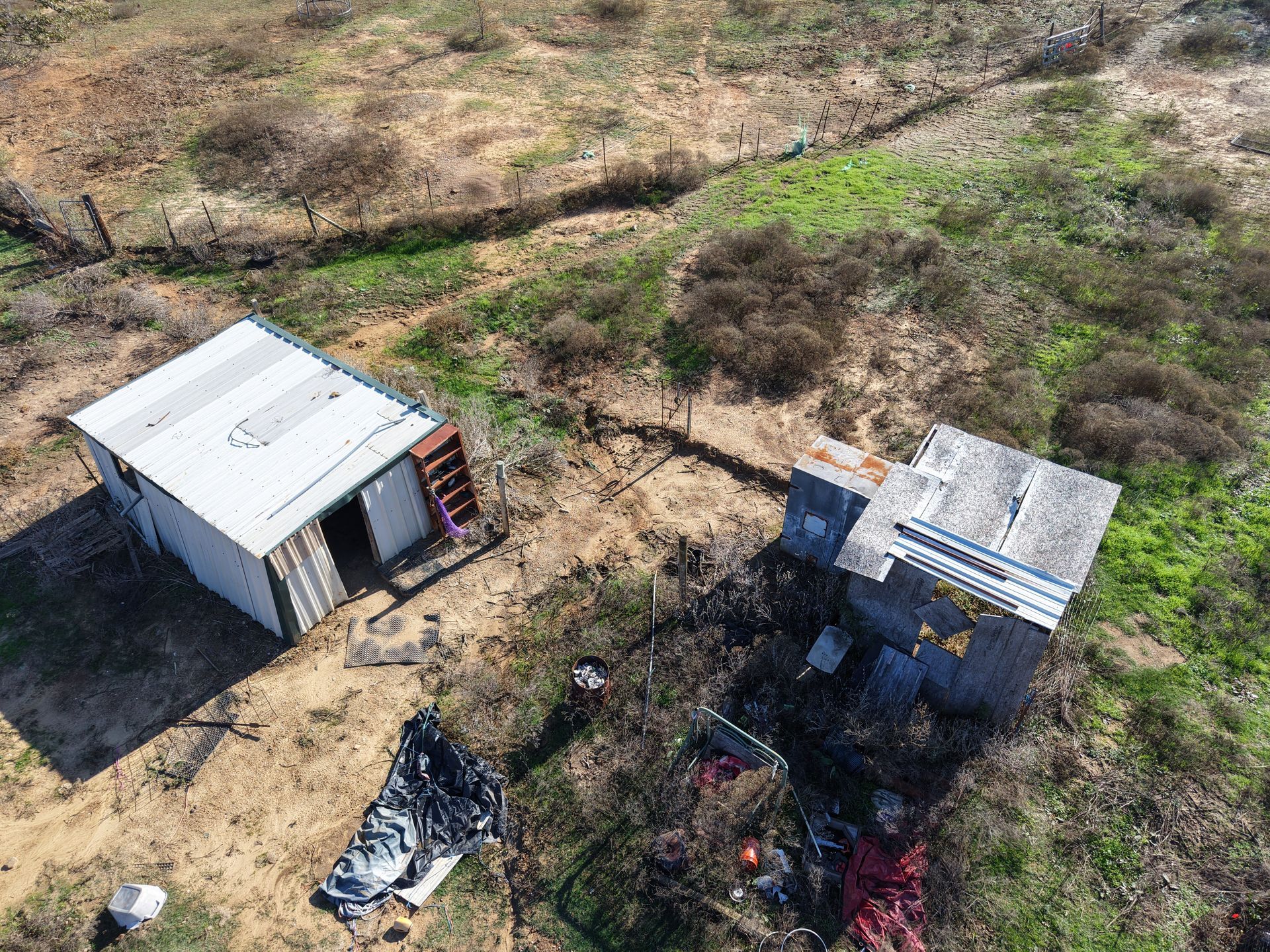 Two small sheds on dry grassland, with scattered debris and a shadowed area nearby.