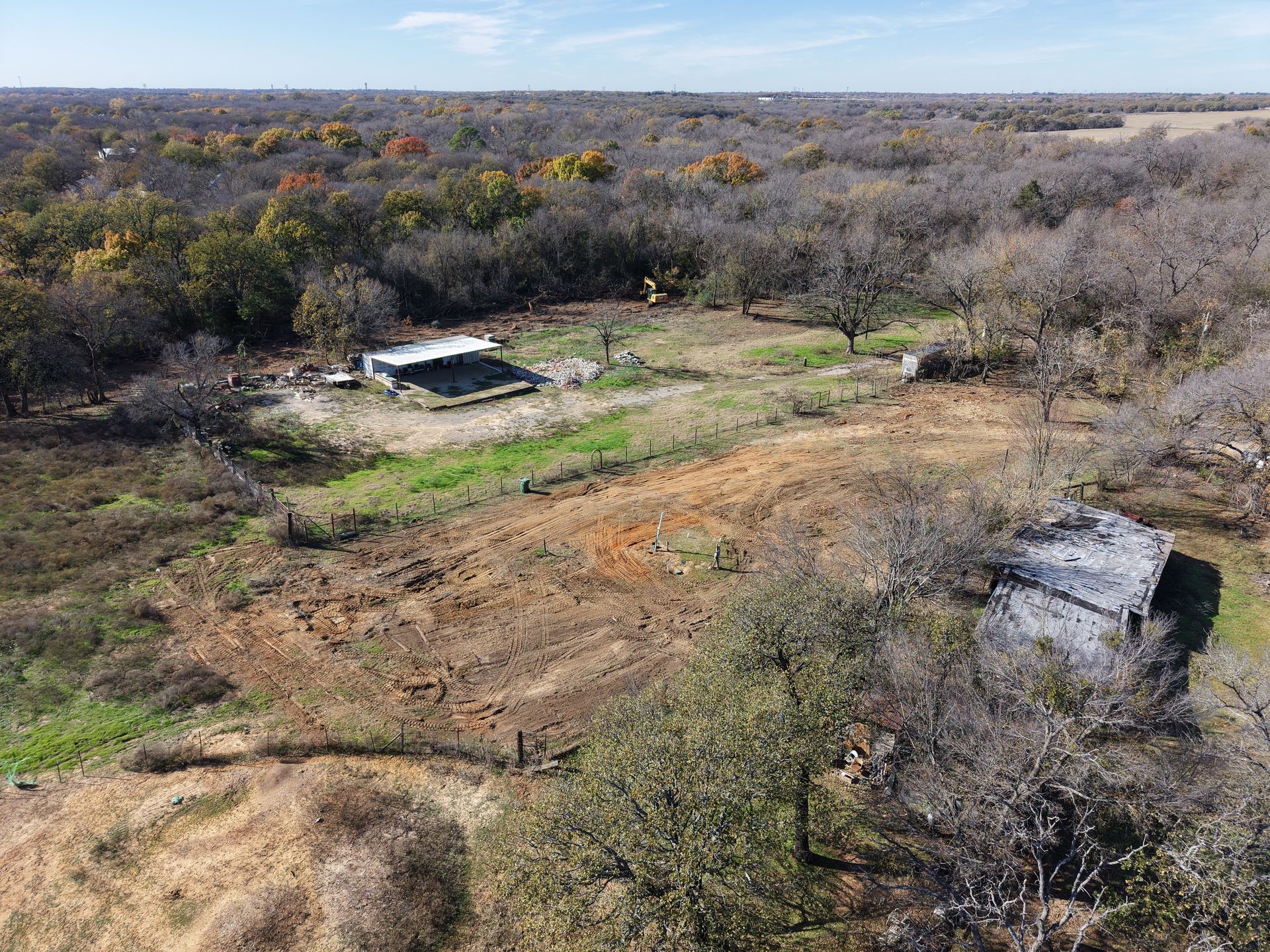 Dry grassy field with scattered shrubs, a small concrete structure, and trees under a clear sky.