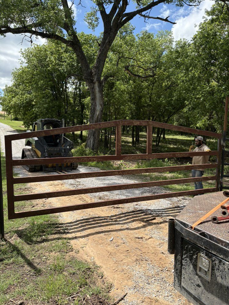 Closed rusty gate blocking a gravel driveway beside trees and a tractor under bright sunlight